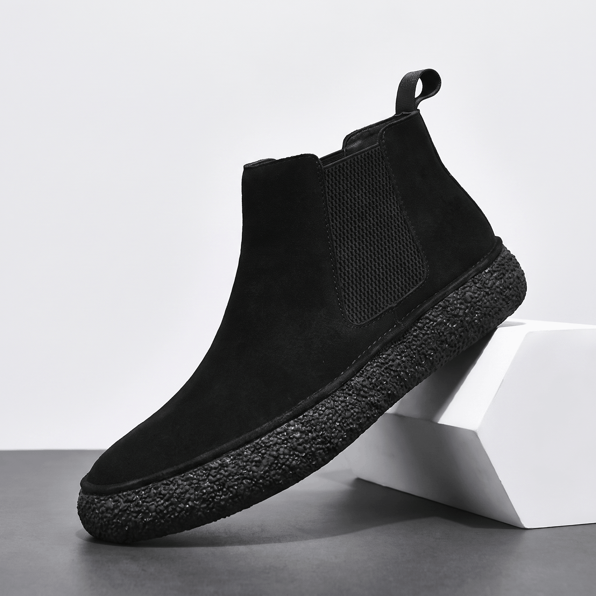 Single black suede Chelsea boot with an elastic side panel, pull tab, and thick crepe sole on a white pedestal.