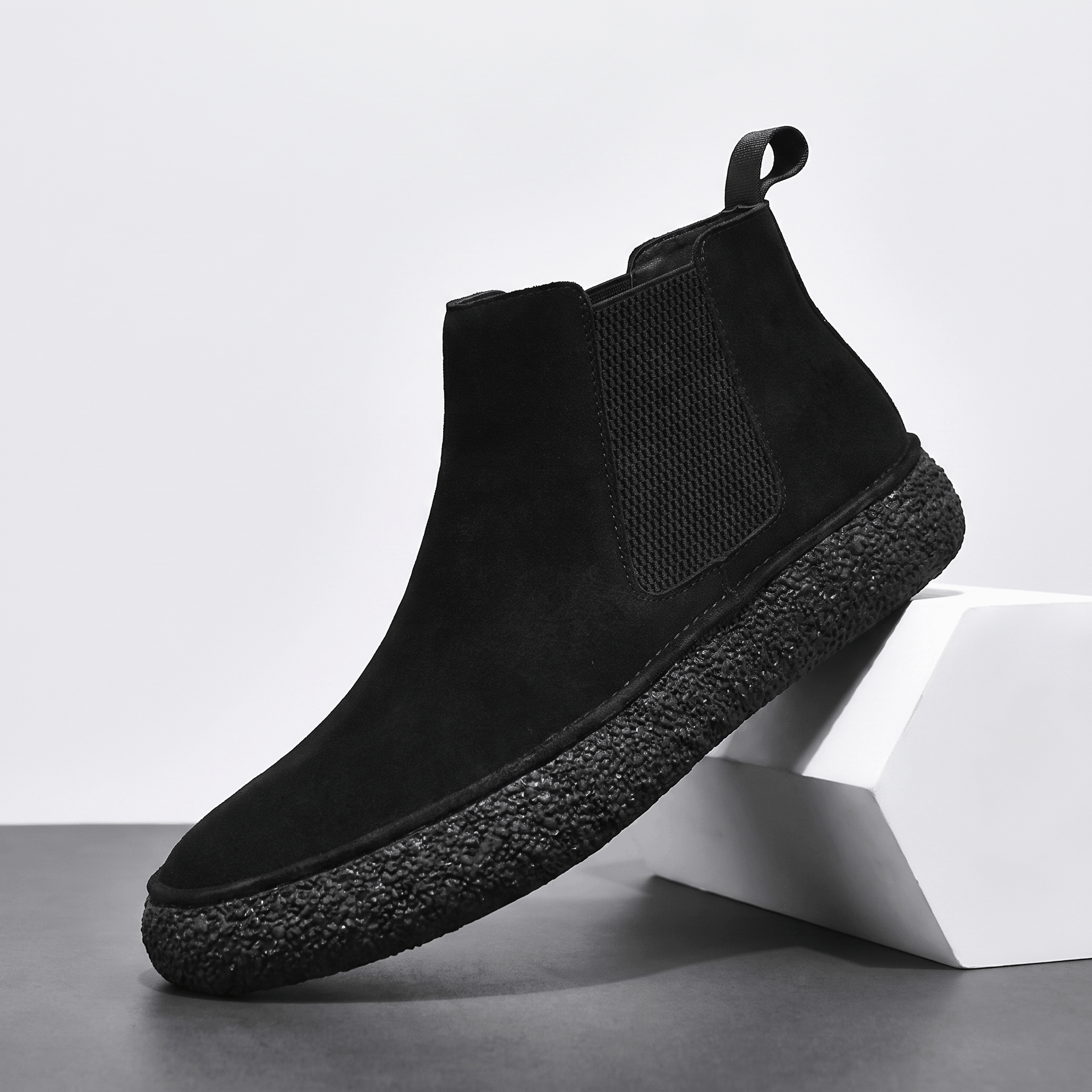 Single black suede Chelsea boot with an elastic side panel, pull tab, and thick crepe sole on a white pedestal.