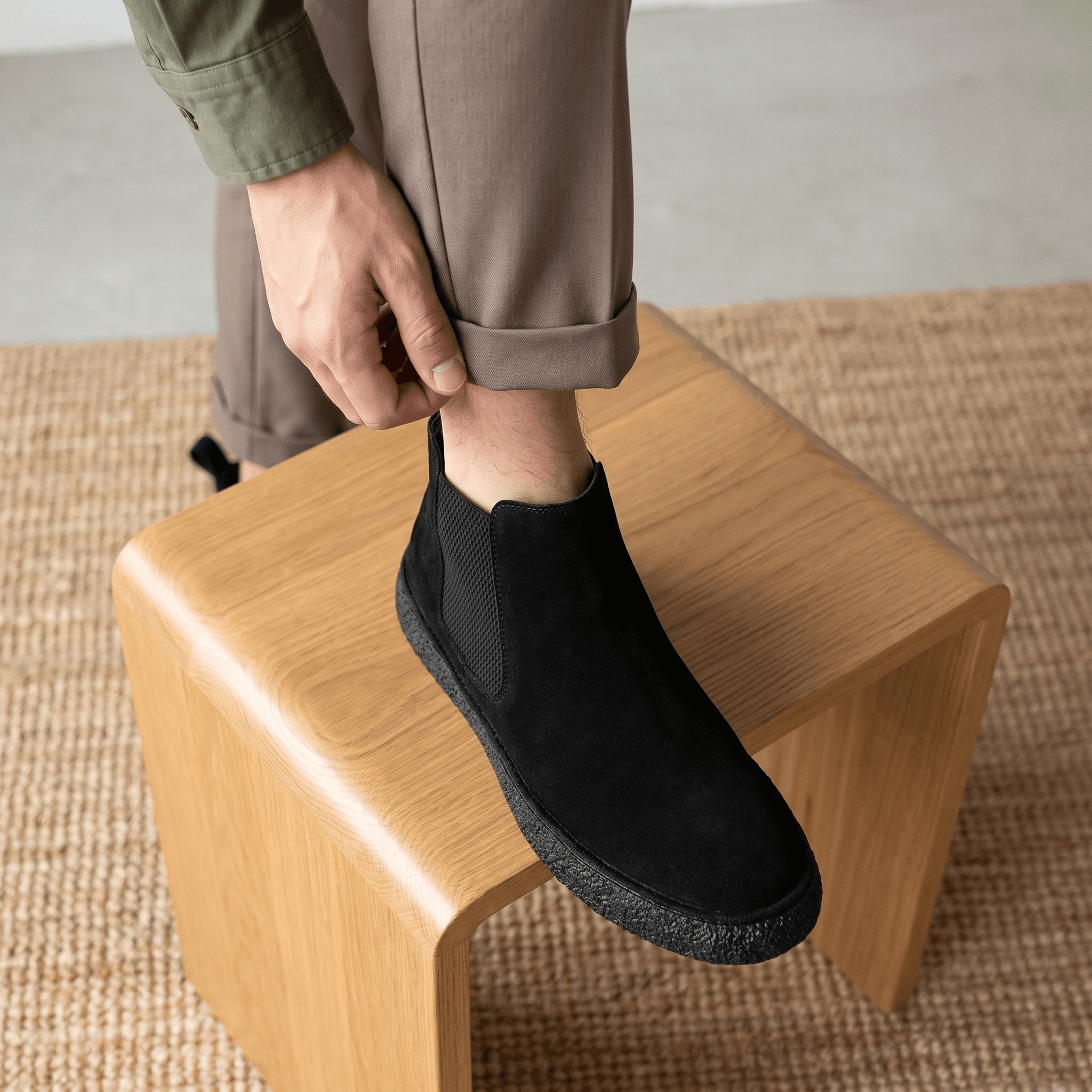 Man seated indoors wearing black suede Chelsea boots with dark trousers and a flat crepe sole.