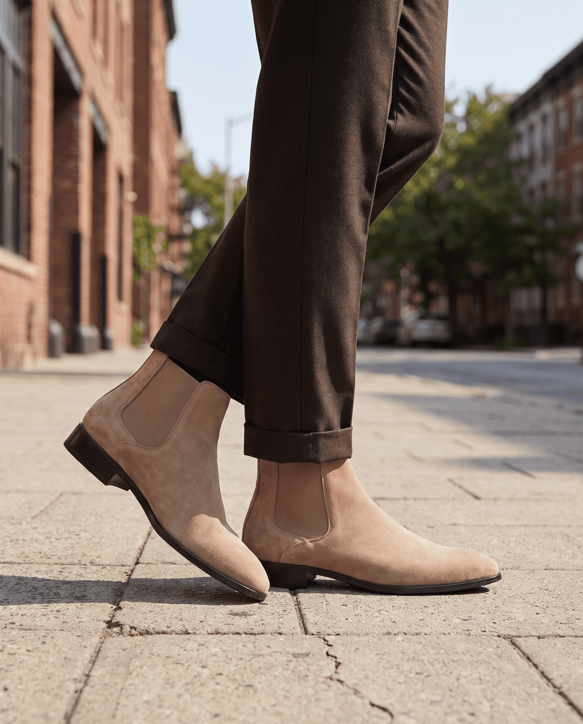 Man in taupe suede boots and dark trousers walking on a brick-lined street
