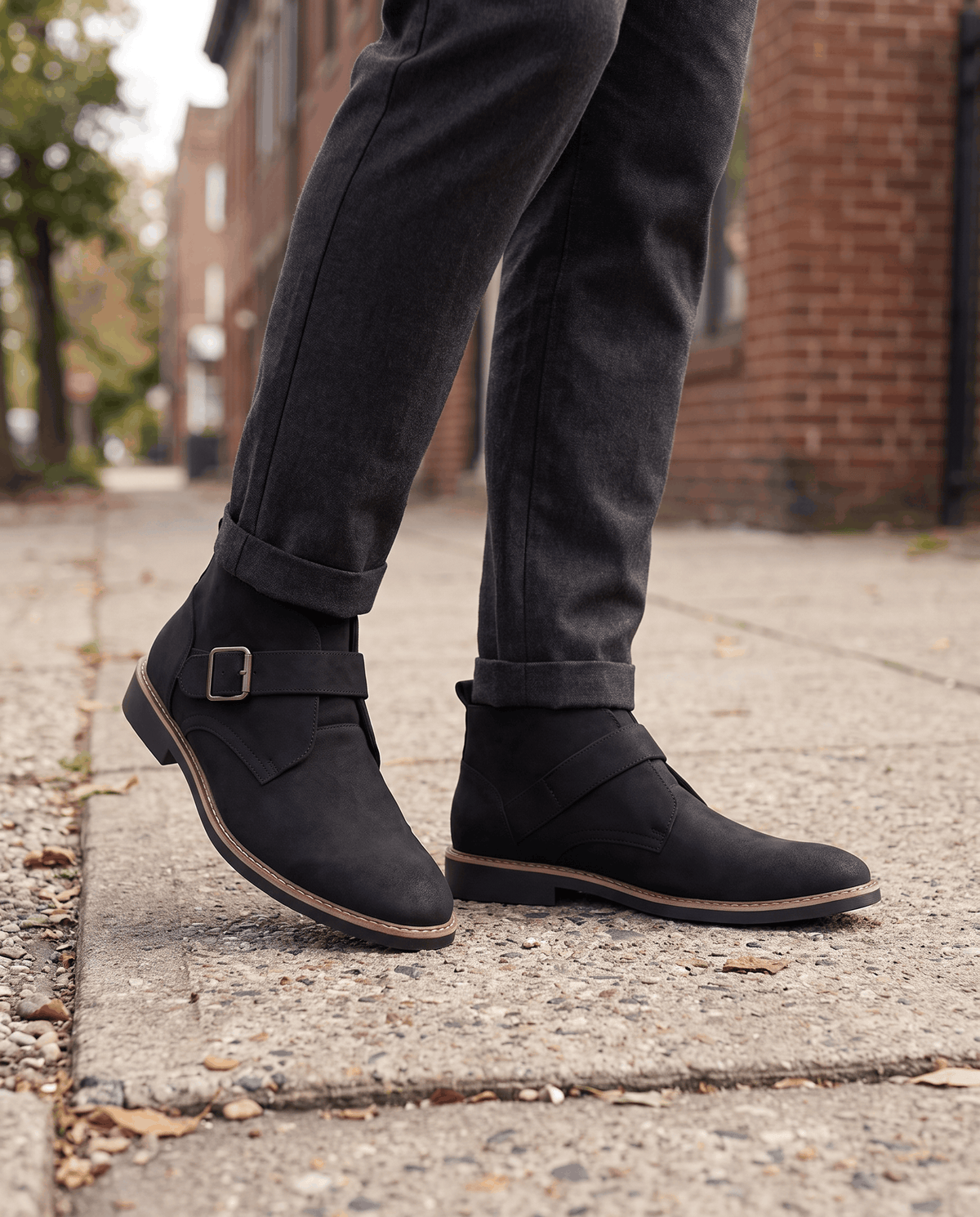 Black suede boots worn with dark grey trousers on a paved street by a brick wall
