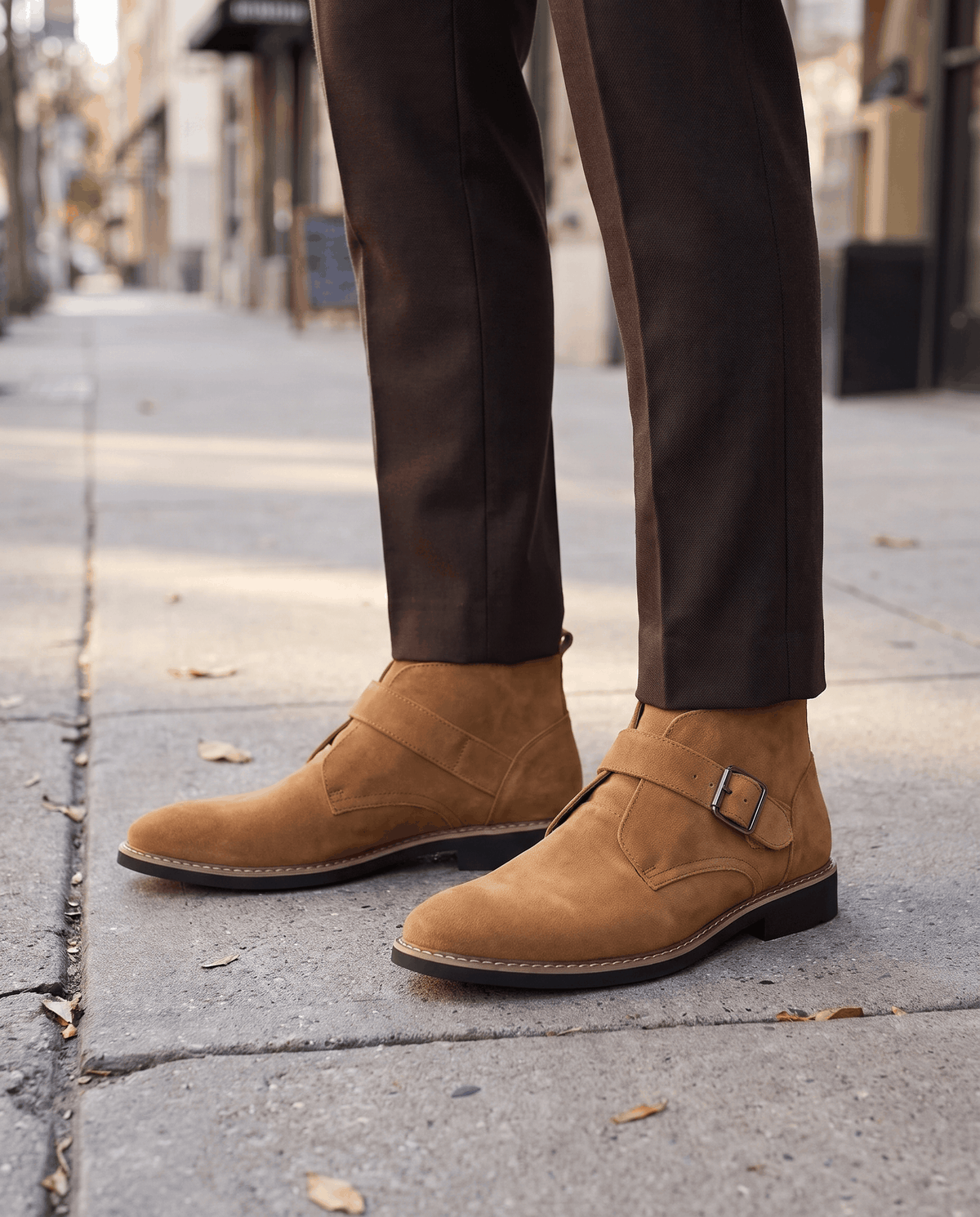 Tan suede buckle boots worn with dark brown trousers on a paved sidewalk