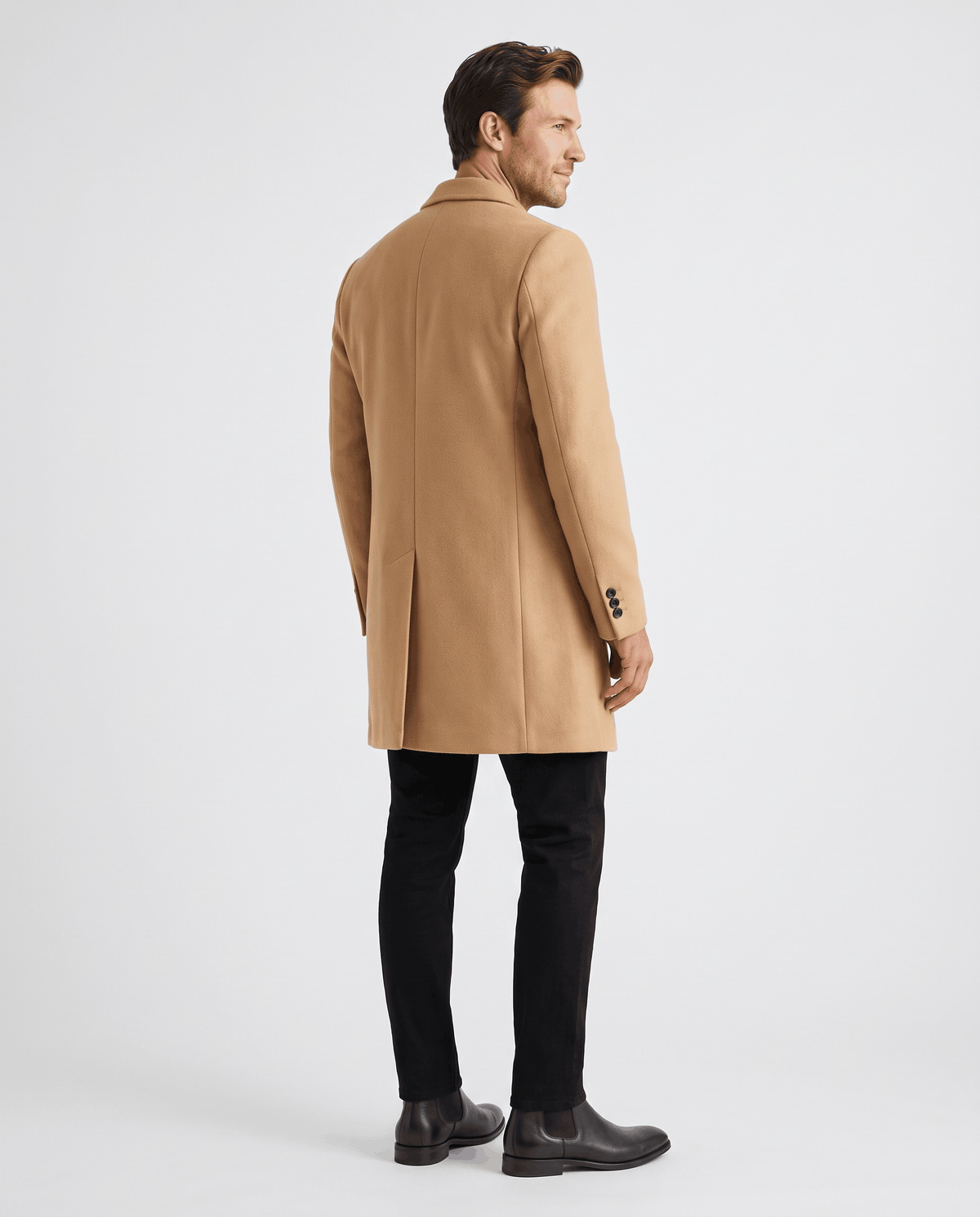 Back view of the khaki overcoat showing the straight back panel, worn with black trousers