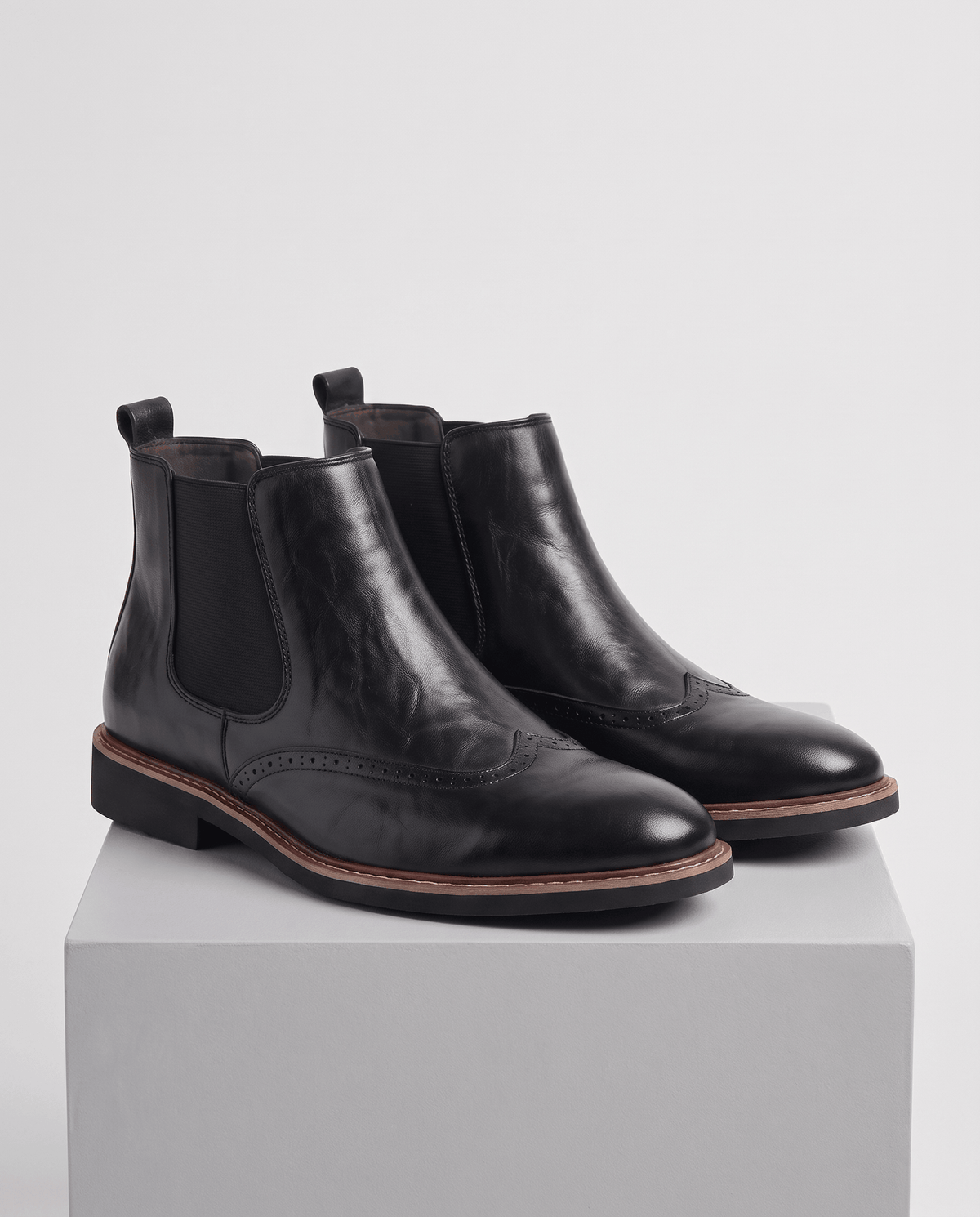 Pair of black wingtip boots with elastic side panels on a white pedestal