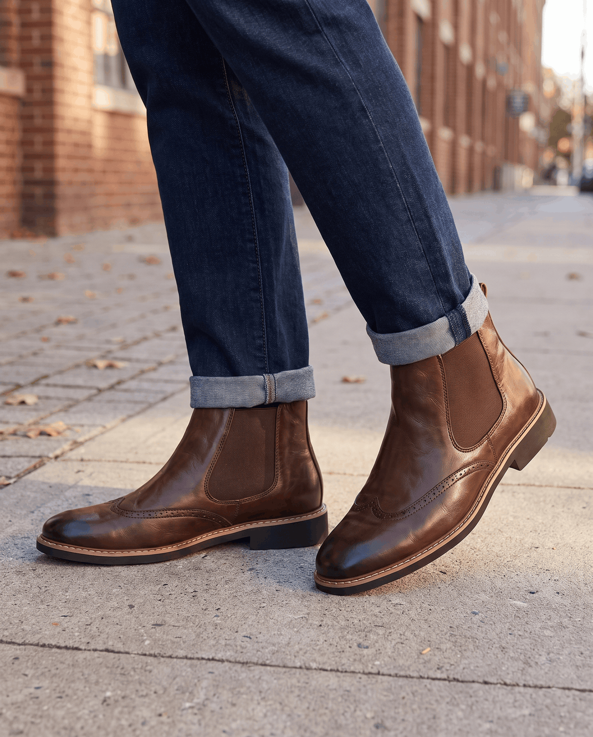 Brown brogue boots worn with rolled dark blue jeans on a paved street