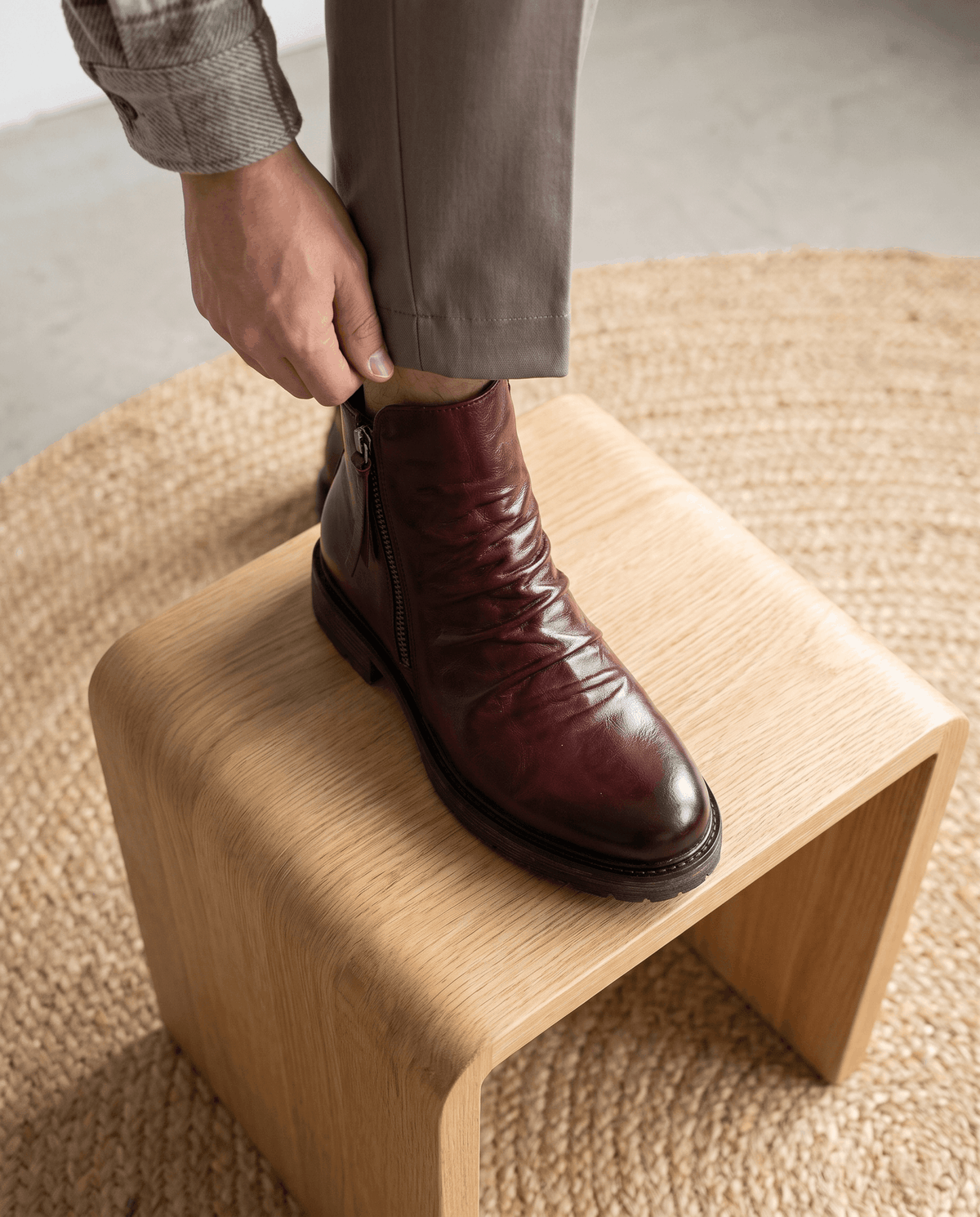 Dark red waxed leather boots with a ruched shaft, side zip, and slip-resistant rubber tread resting on a wood stool.