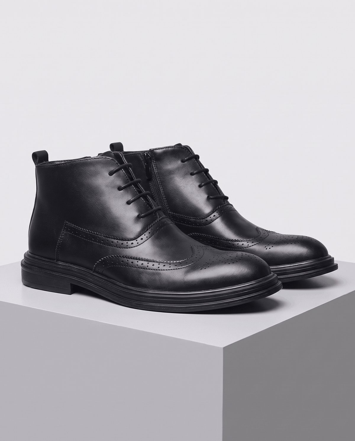 Black brogue ankle boots placed side by side on a gray block