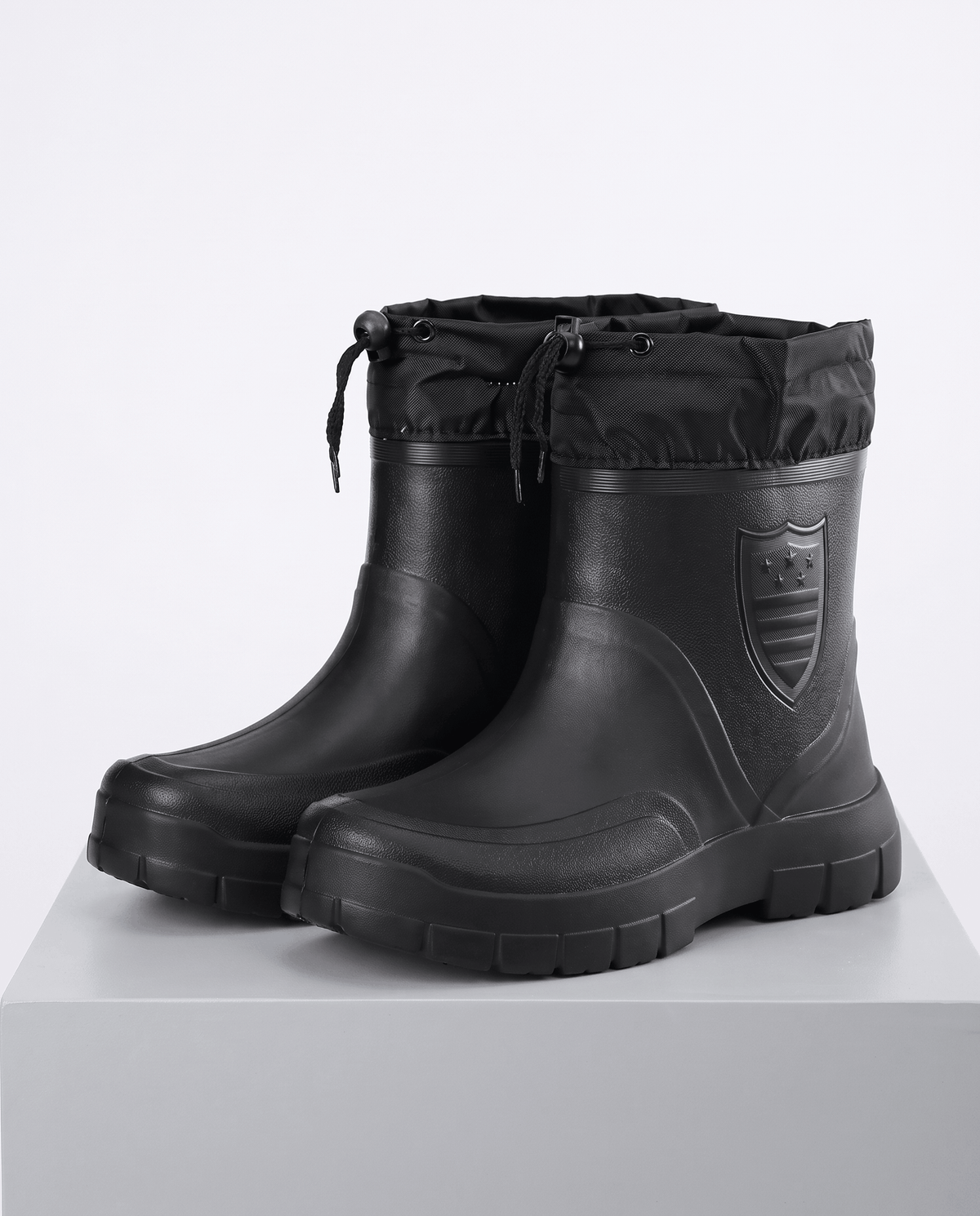 Black mid-calf rain boots with drawstring collars and treaded soles on a gray plinth