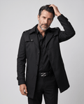 Mid-length single-breasted trench coat in black, worn buttoned over a dark shirt, model hand raised
