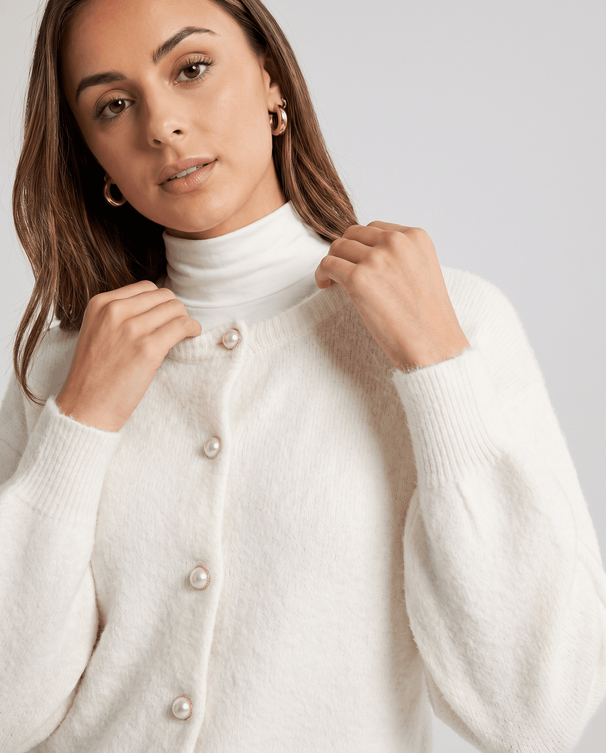 Model framing the crew neck of the white cardigan, showing its pearl-effect buttons