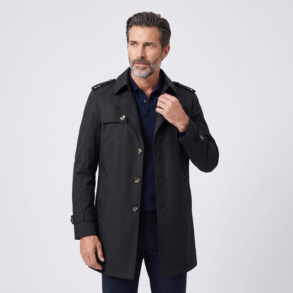 Model in black trench coat buttoned over navy shirt, hand at collar, front view