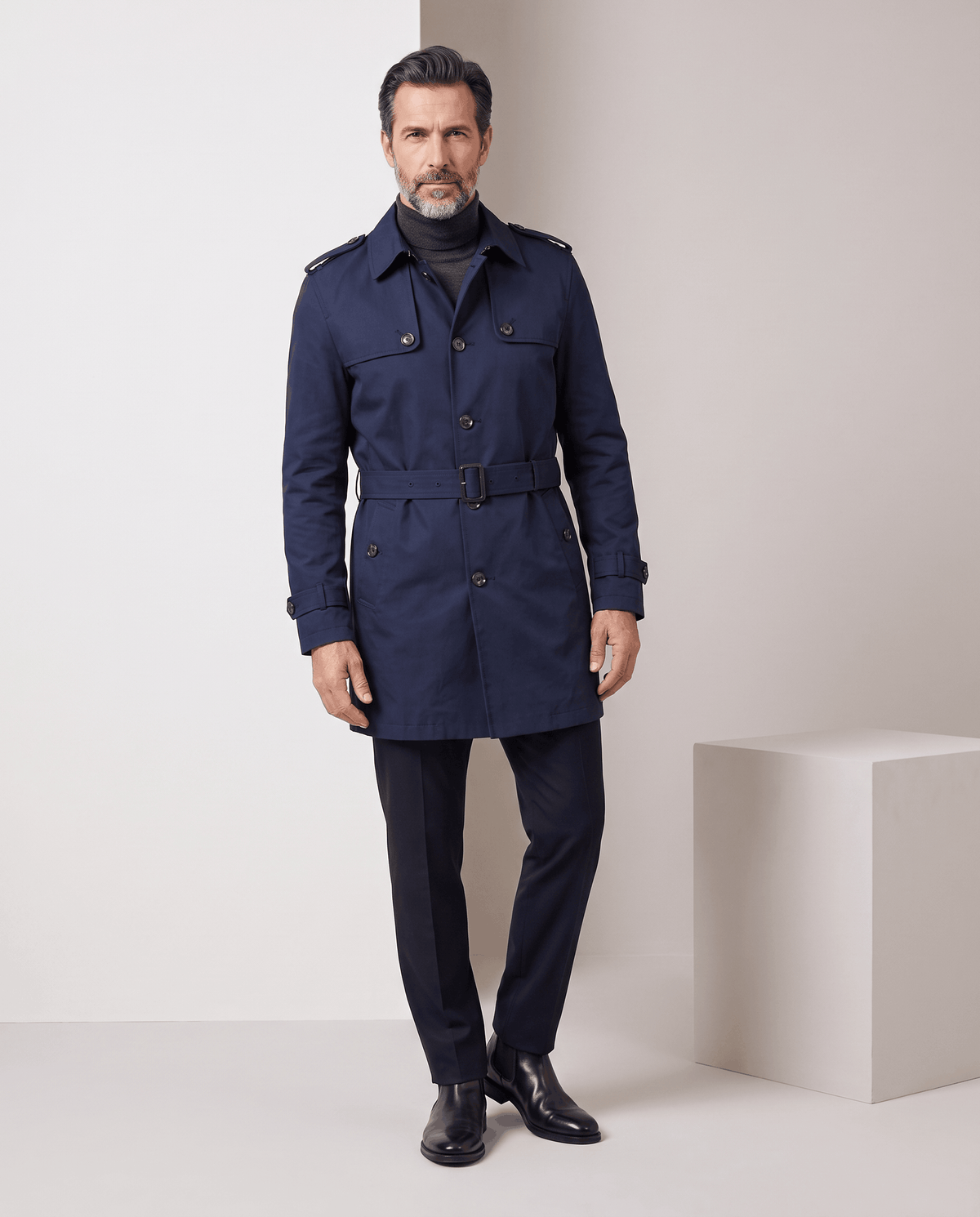 Full-length navy trench coat with belt, styled with dark trousers and studio backdrop