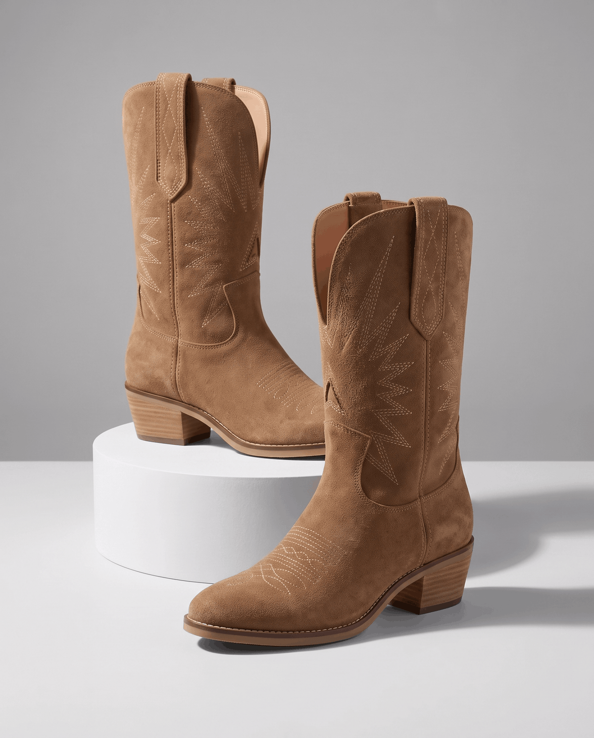 Two tan suede boots on a white pedestal against a grey studio backdrop