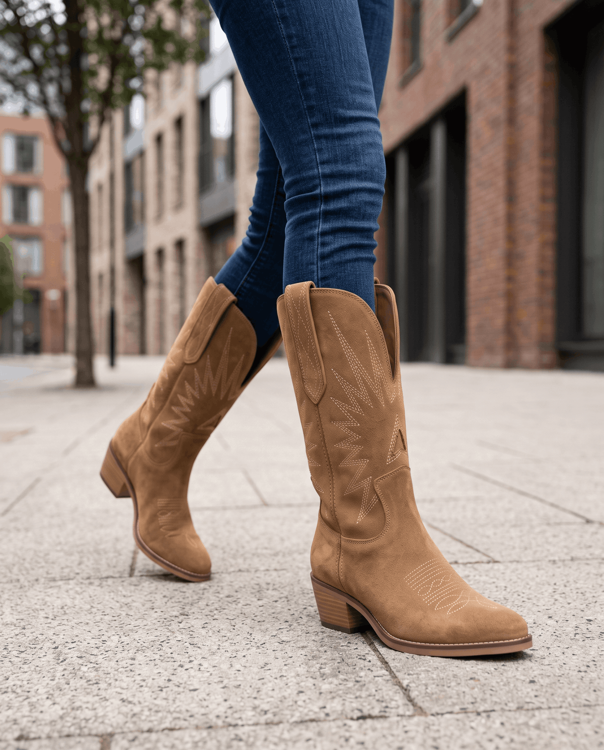 Legs in blue jeans and stitched suede boots walking along a city sidewalk