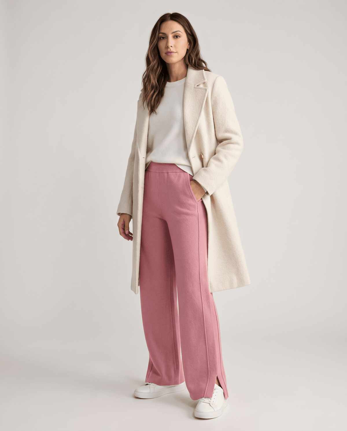 Model in long cream coat and white top wearing pink full-length knit pants