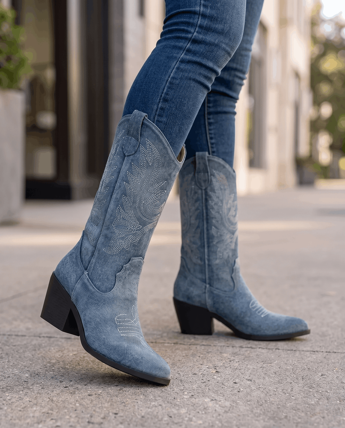 Close view of legs in blue jeans and blue stitched boots with black heels on a sidewalk