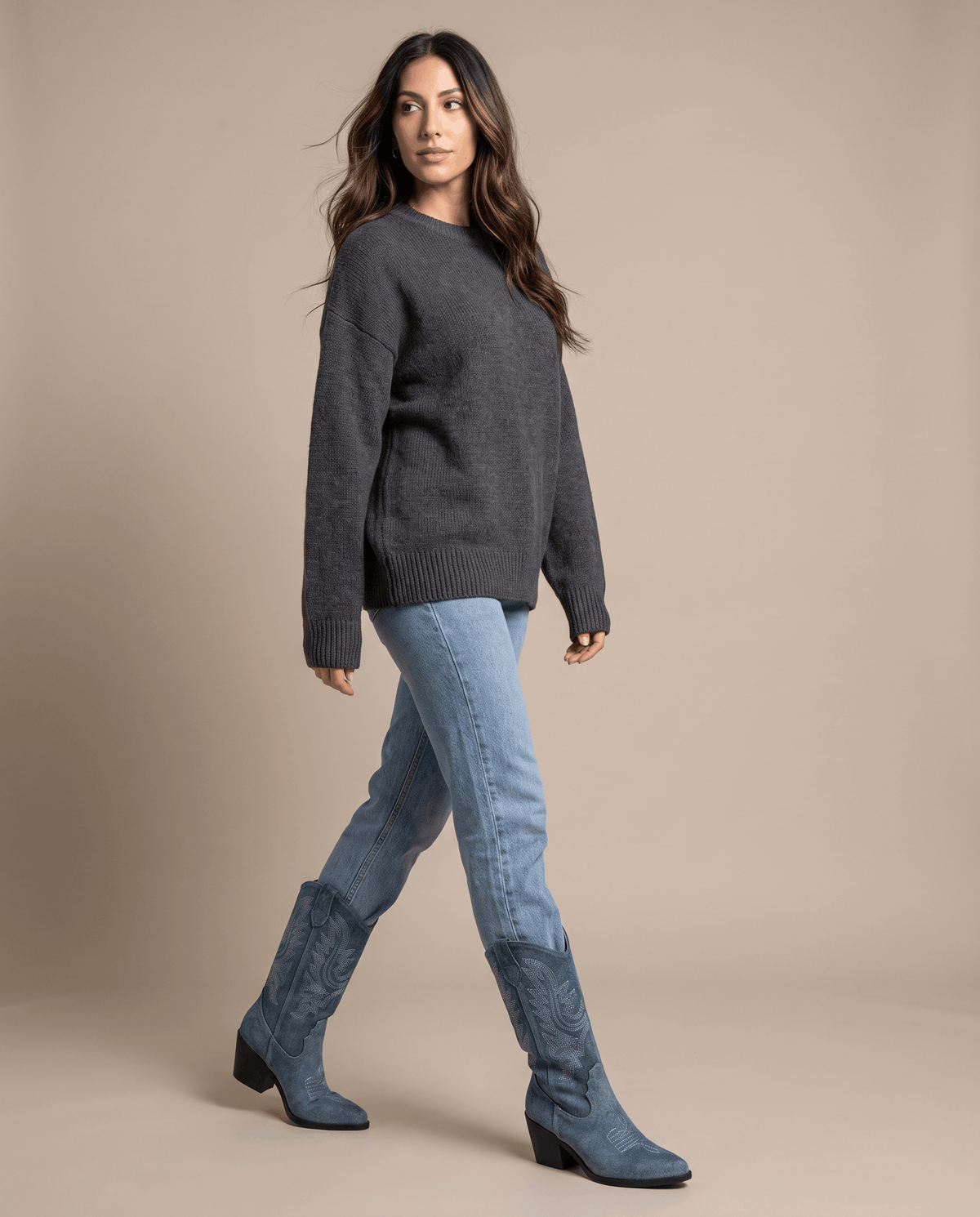 Woman in dark knit sweater and jeans walking in blue stitched boots against a tan backdrop