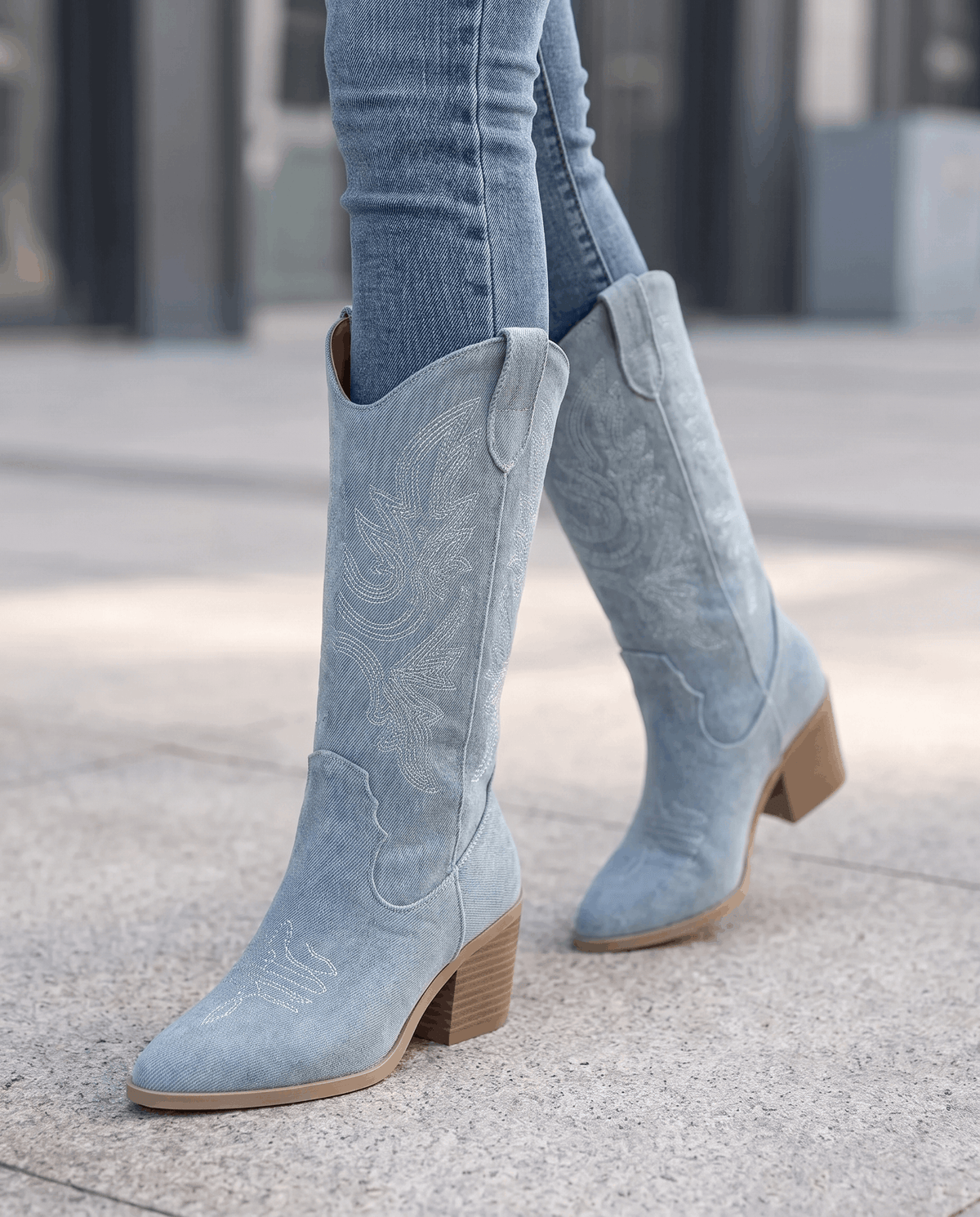 Legs in blue jeans and light blue stitched boots walking along a city sidewalk