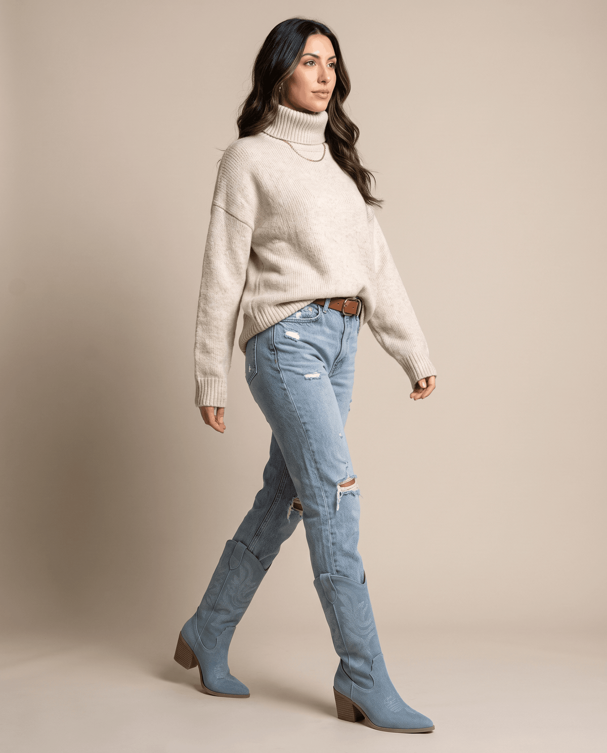Woman in cream knit sweater and jeans walking in light blue Western boots against a tan backdrop