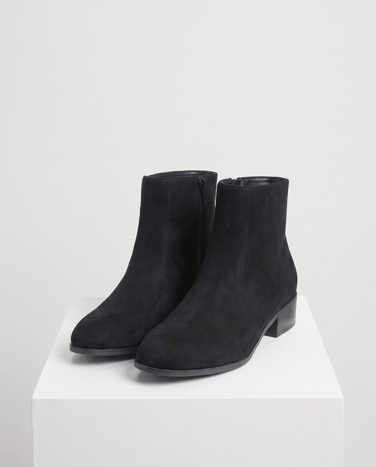 Black suede-look ankle boots styled on a white pedestal