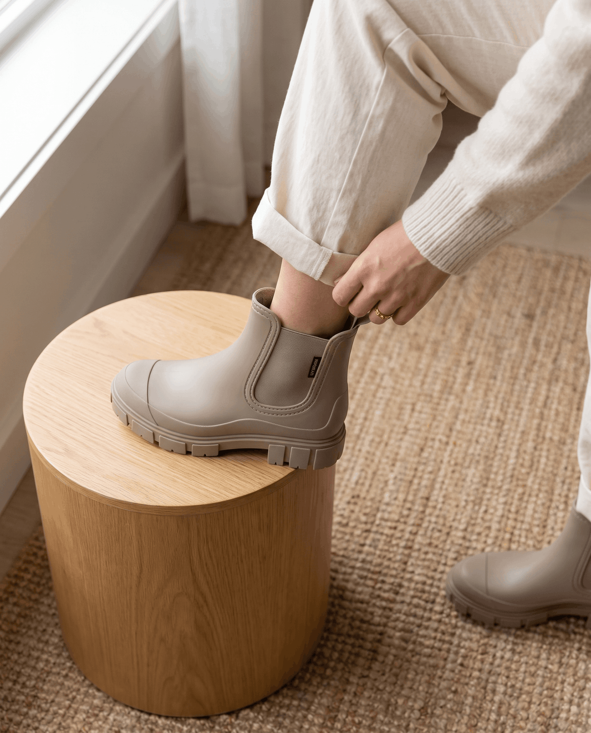 Person putting on taupe Chelsea boots while seated beside a wooden stool.