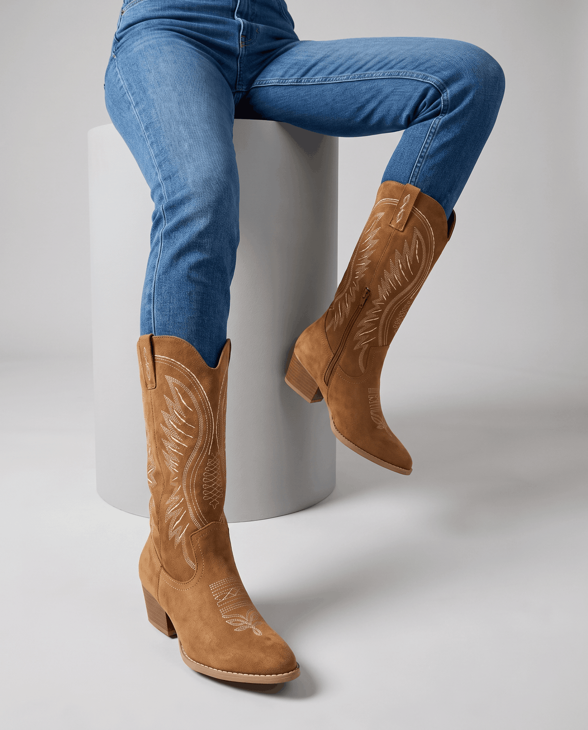 Seated model with blue jeans tucked into brown embroidered boots on a white block