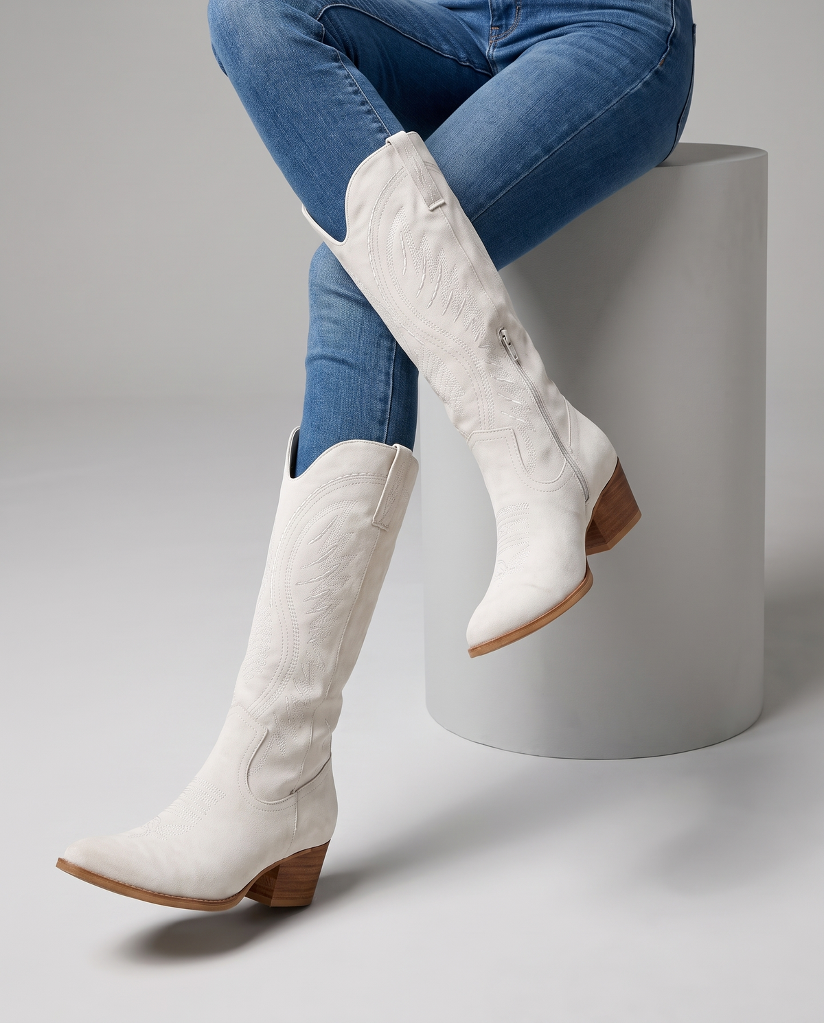 Seated model with blue jeans tucked into white embroidered boots on a white block
