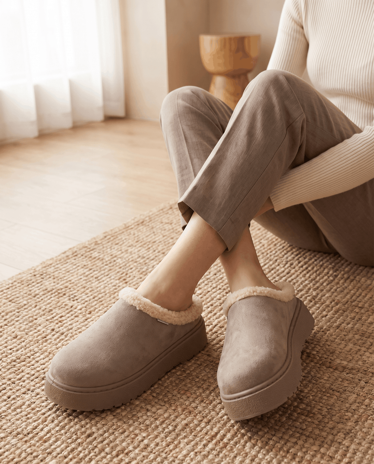 Person wearing taupe fleece-lined slippers with shearling-look cuffs while seated indoors.