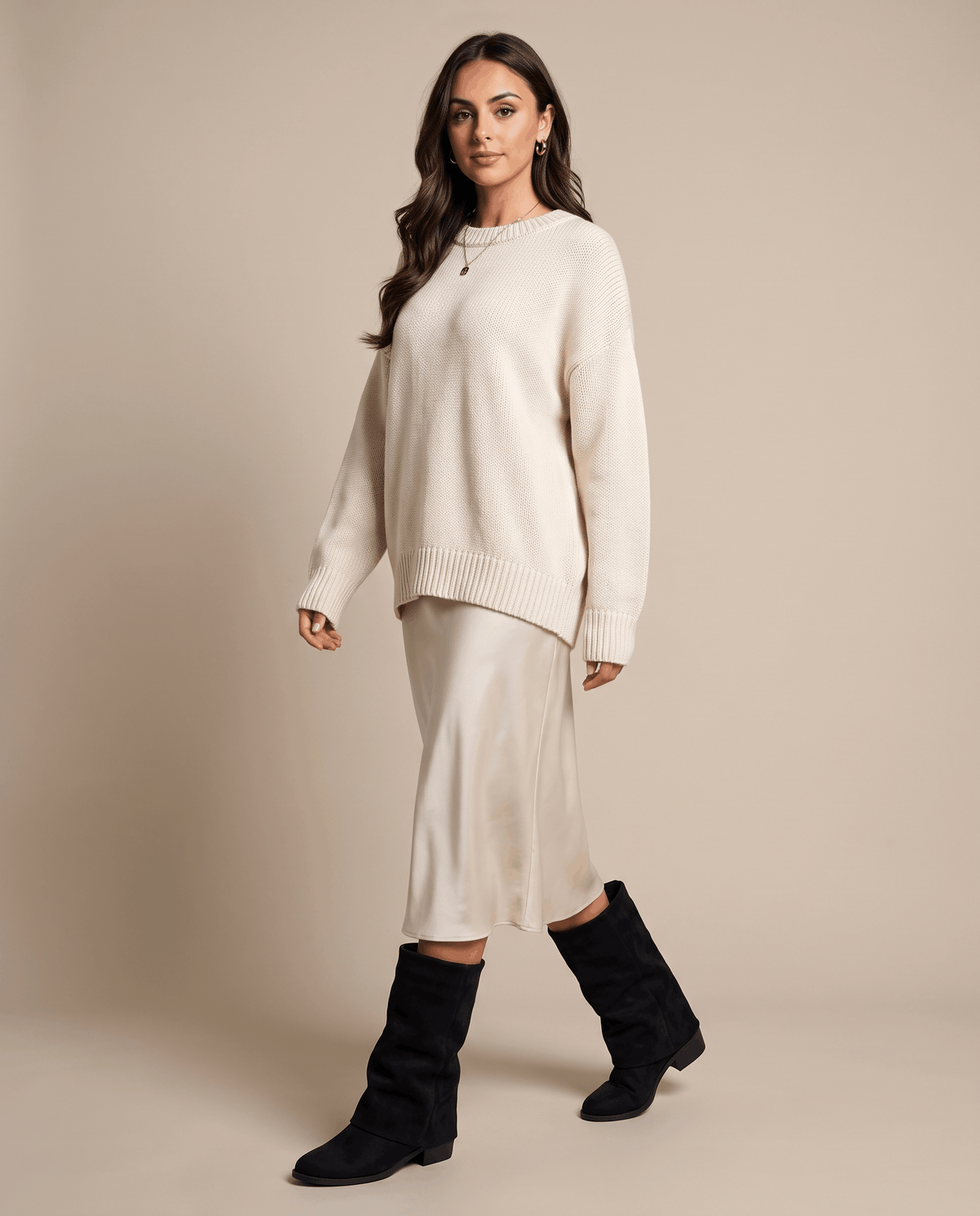 Woman in a cream sweater and satin midi skirt walking in black fold-over boots