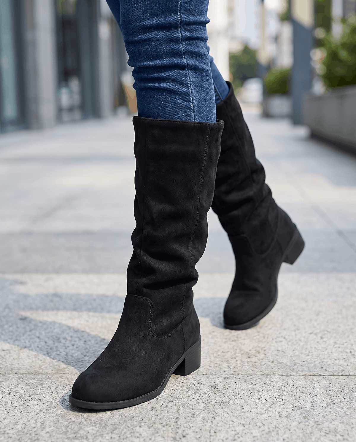 Black suede boots worn walking on a city sidewalk with blue jeans