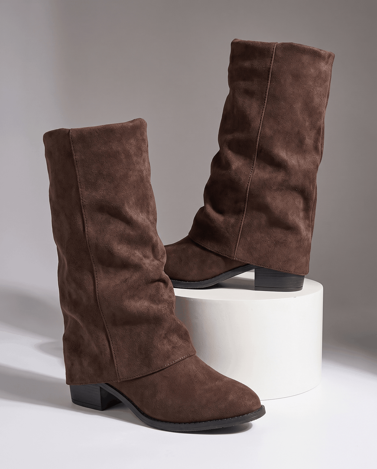 Brown fold-over boots displayed on a white pedestal against a grey backdrop
