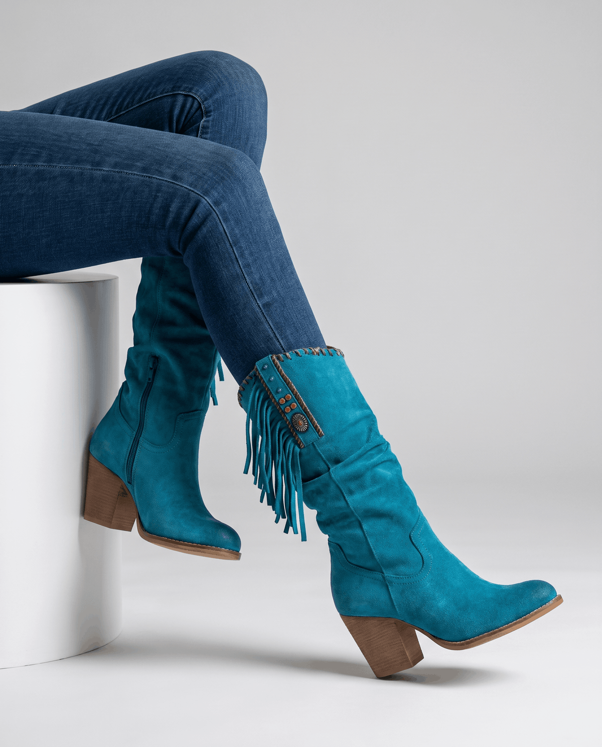 Seated model with tucked skinny jeans wearing blue fringe boots on a white block