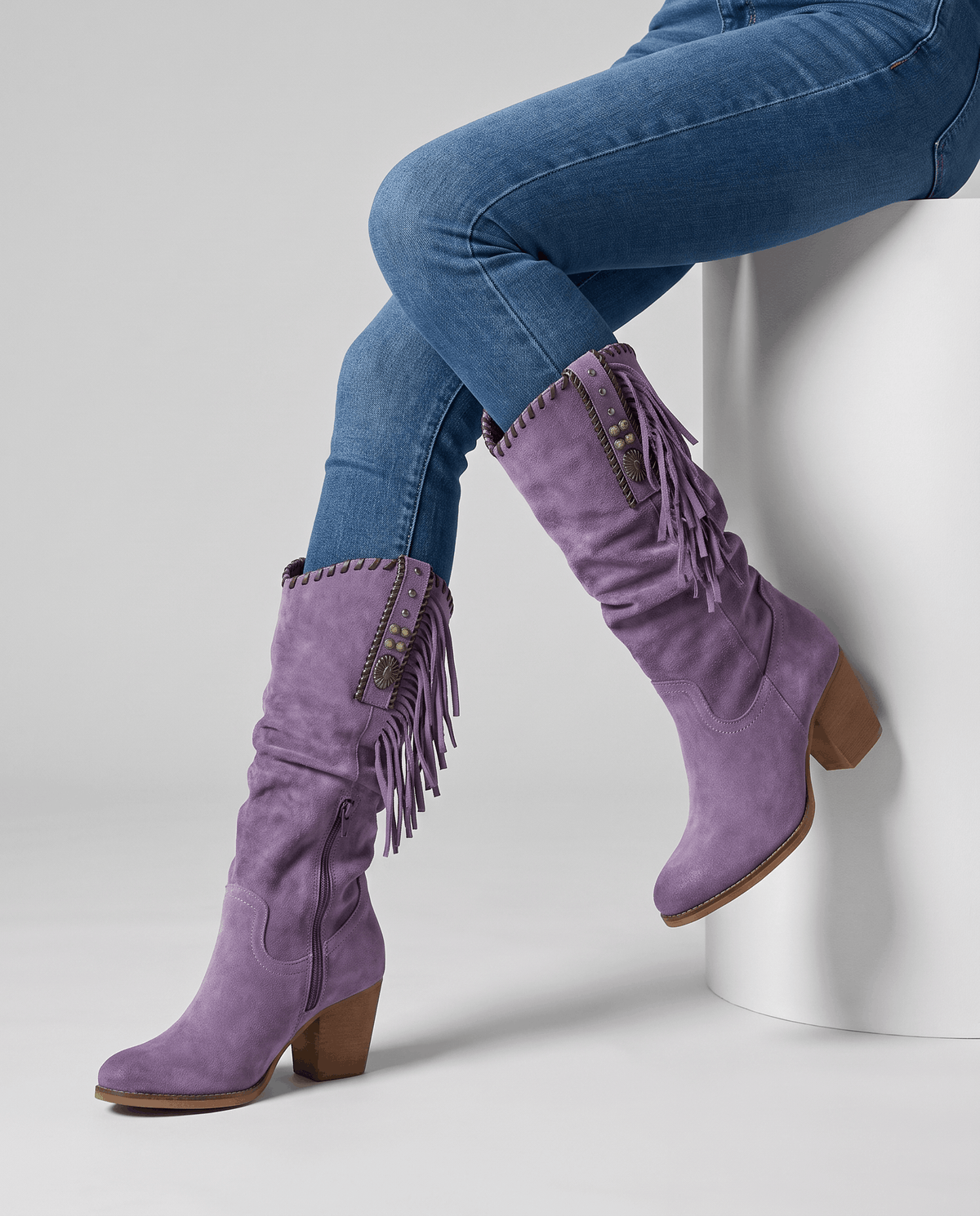 Seated model in blue jeans wearing purple fringe boots on a white block