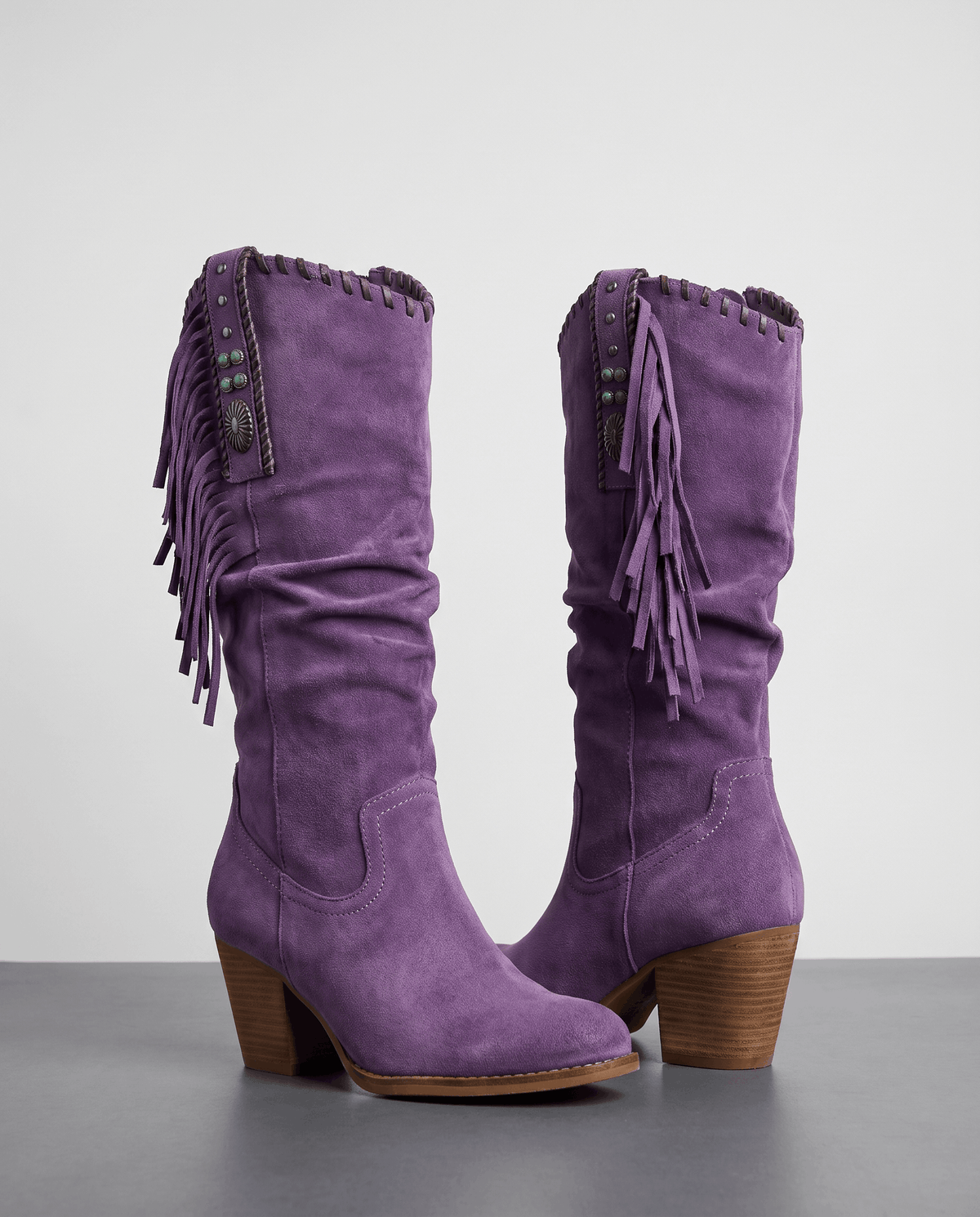 Pair of purple slouchy fringe boots with concho straps on a grey studio floor
