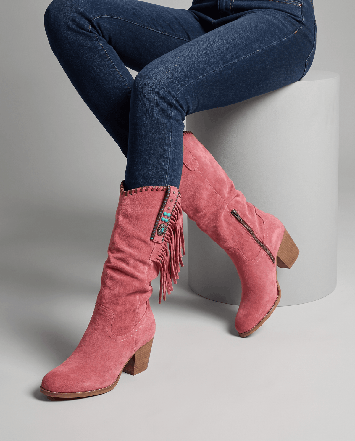 Seated model with blue jeans tucked into red slouchy fringe boots on a white block
