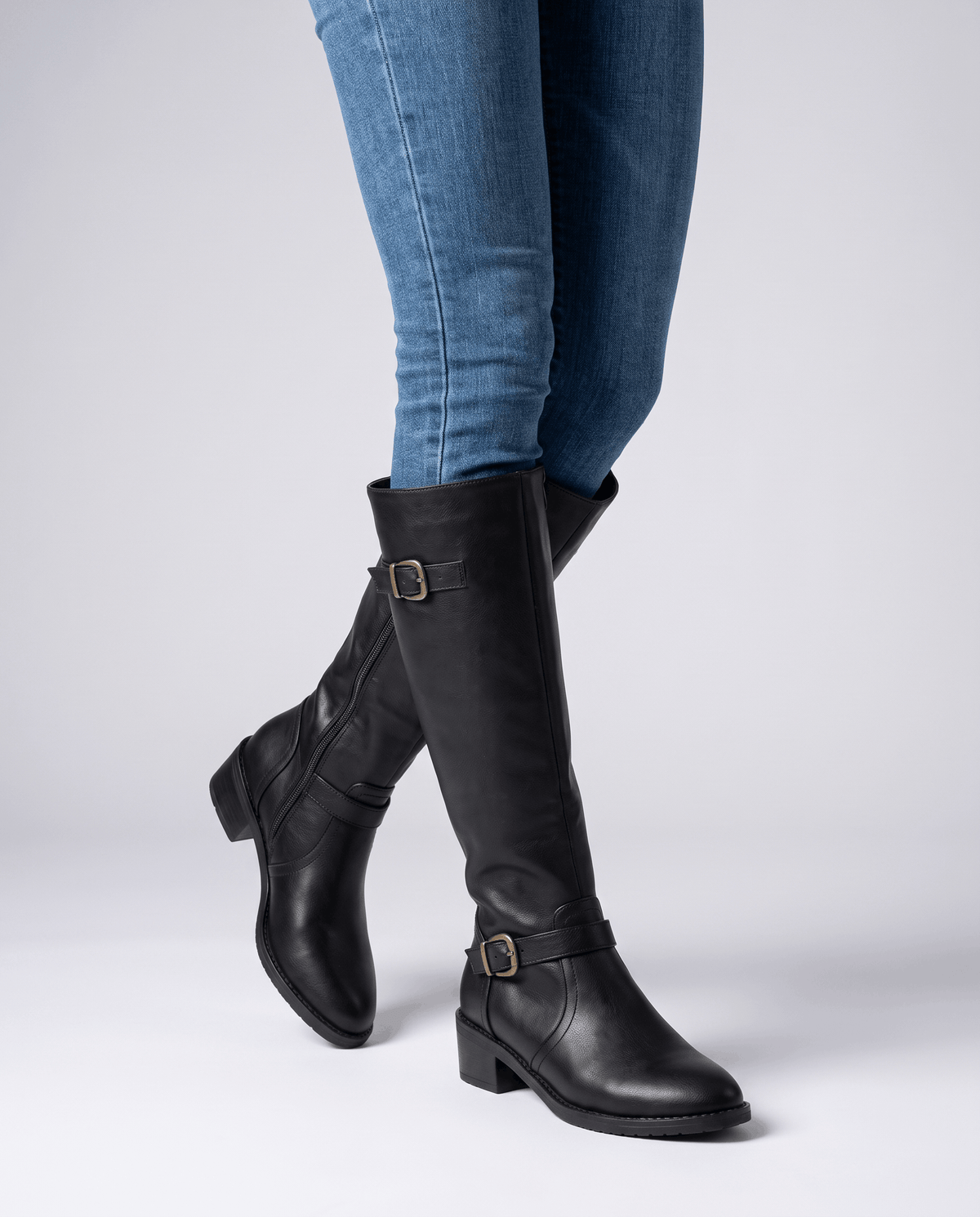Black knee-high riding boots with antique brass buckles, a structured shaft, and a stacked block heel worn with jeans.