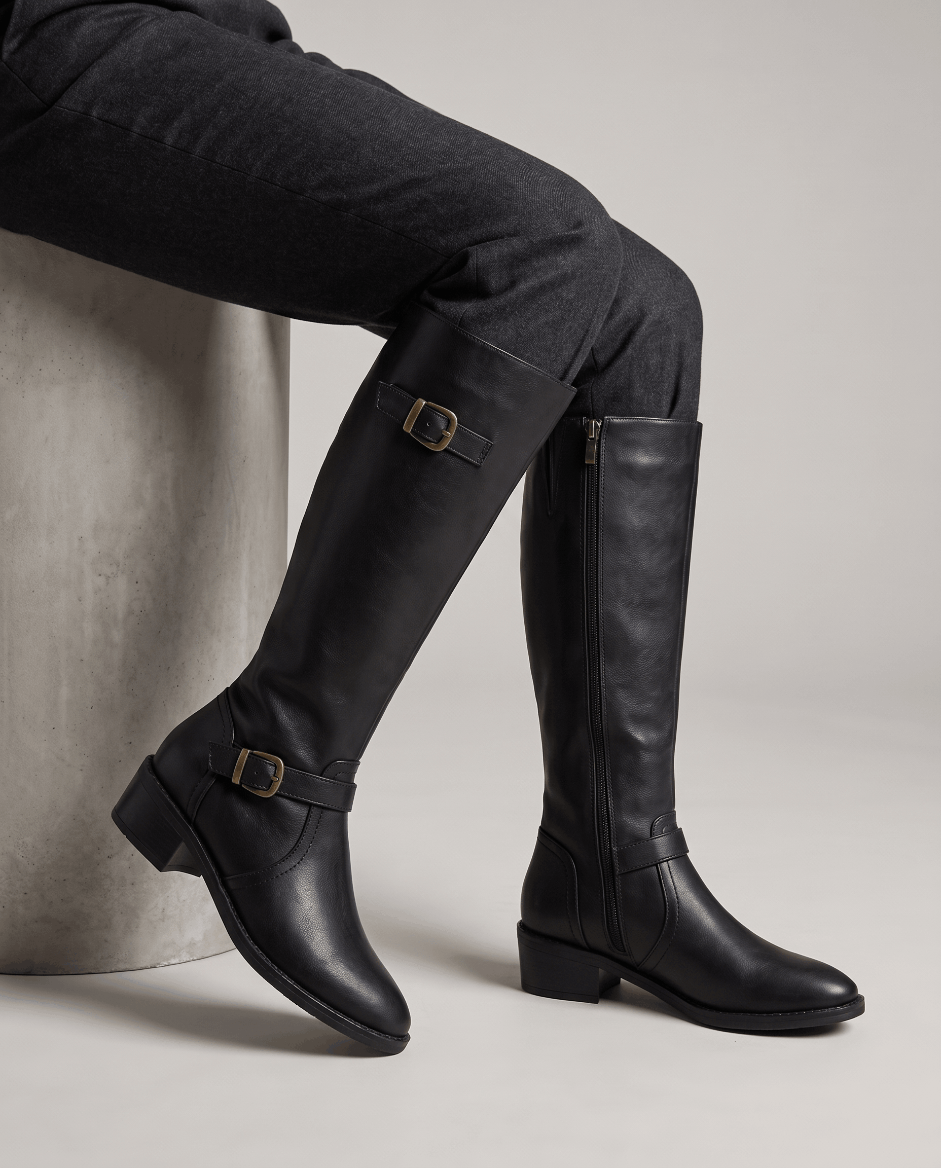 Black knee-high riding boots with antique brass buckles, structured shafts, and stacked block heels worn over dark pants.