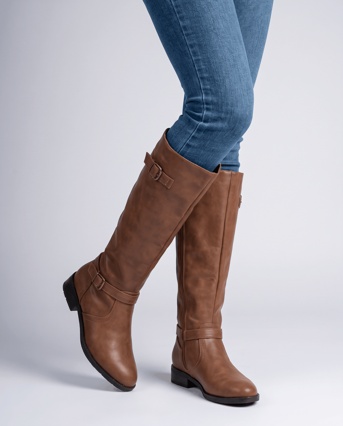 Brown knee-high riding boots with antique brass buckle straps, a structured shaft, and a low stacked block heel.