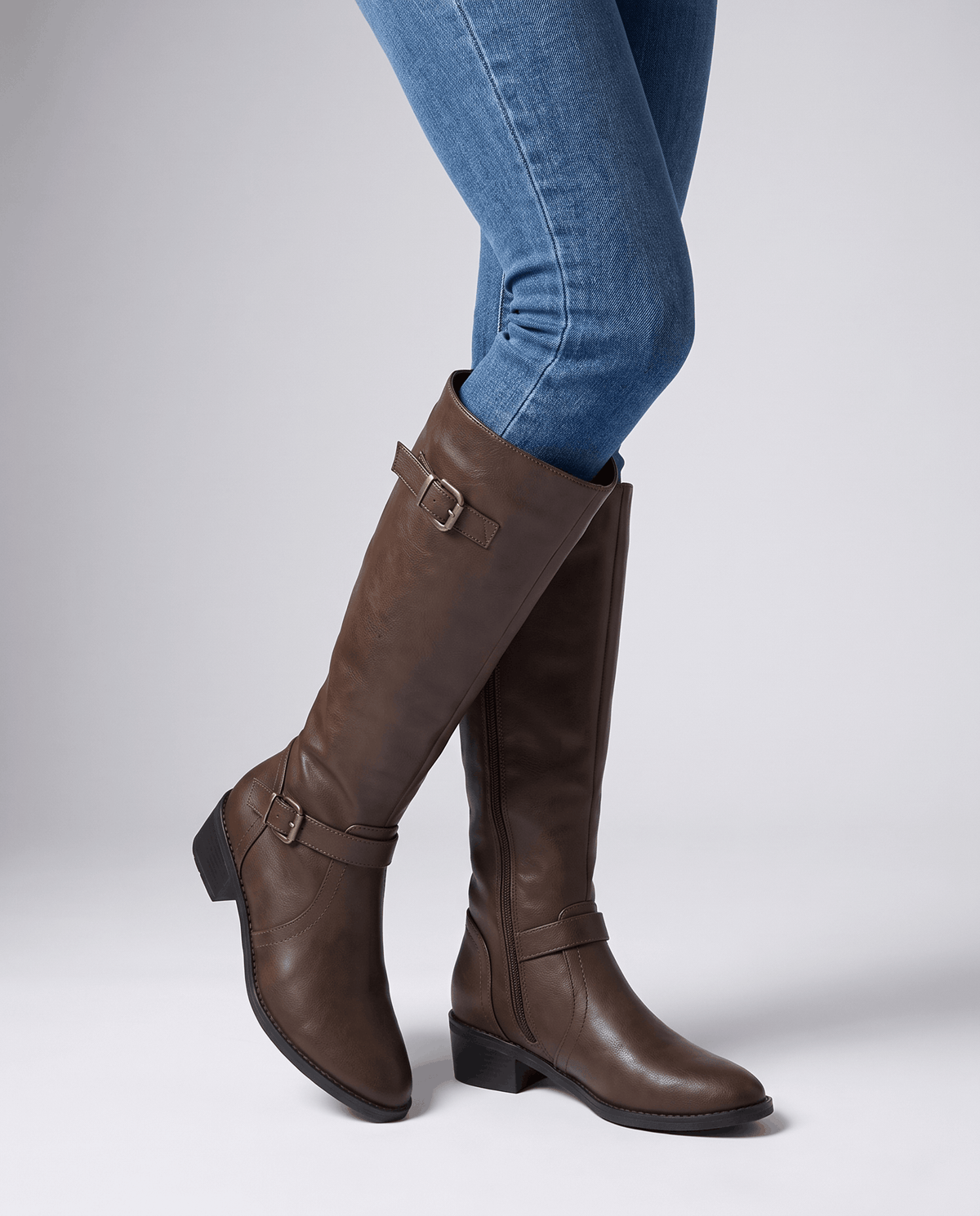 Dark brown knee-high riding boots with antique brass buckles, a structured shaft, and a stacked block heel worn with jeans.
