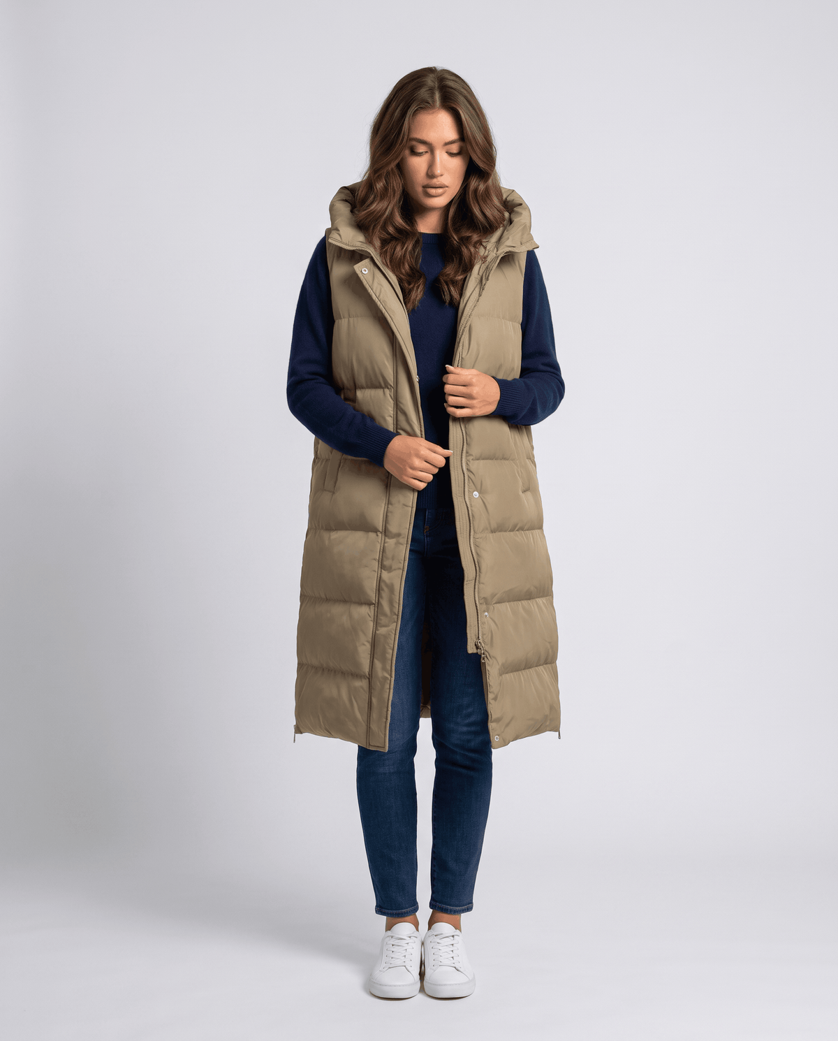 Khaki long quilted hooded vest worn open over a navy sweater and jeans