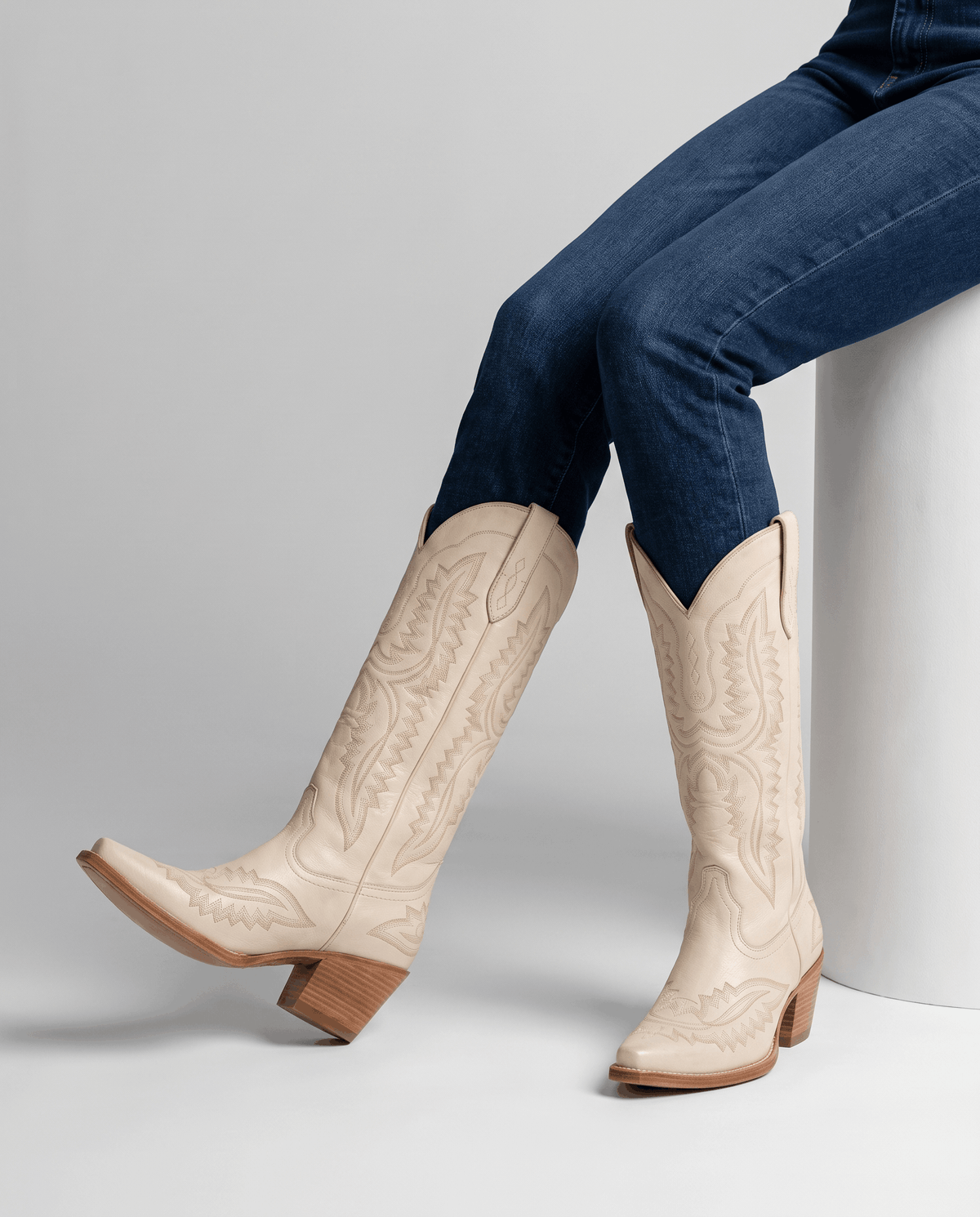 Seated model with blue jeans tucked into cream mid-calf boots with stacked heels
