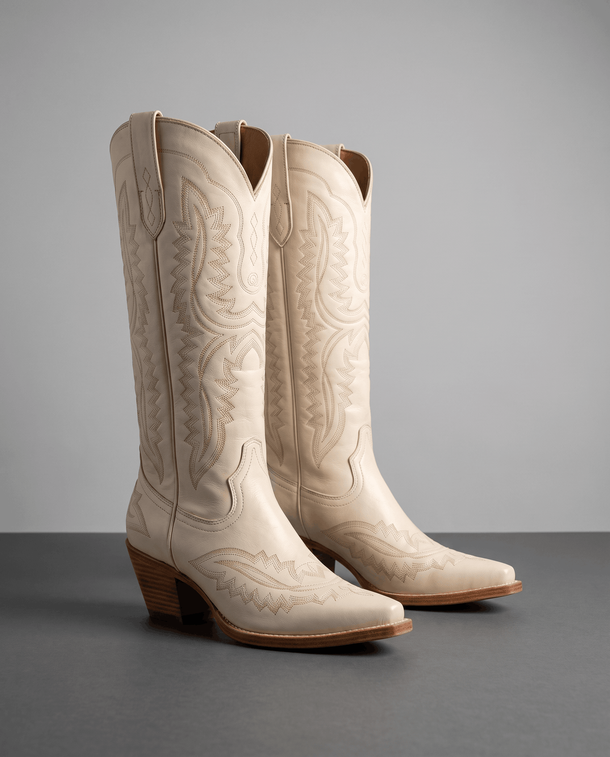 Pair of cream embroidered cowboy boots with tonal stitching on a grey studio floor