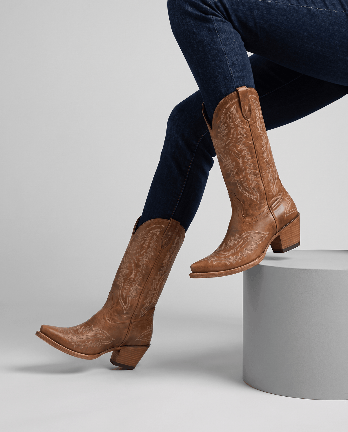 Dark brown boots worn with tucked jeans while seated on a white studio block