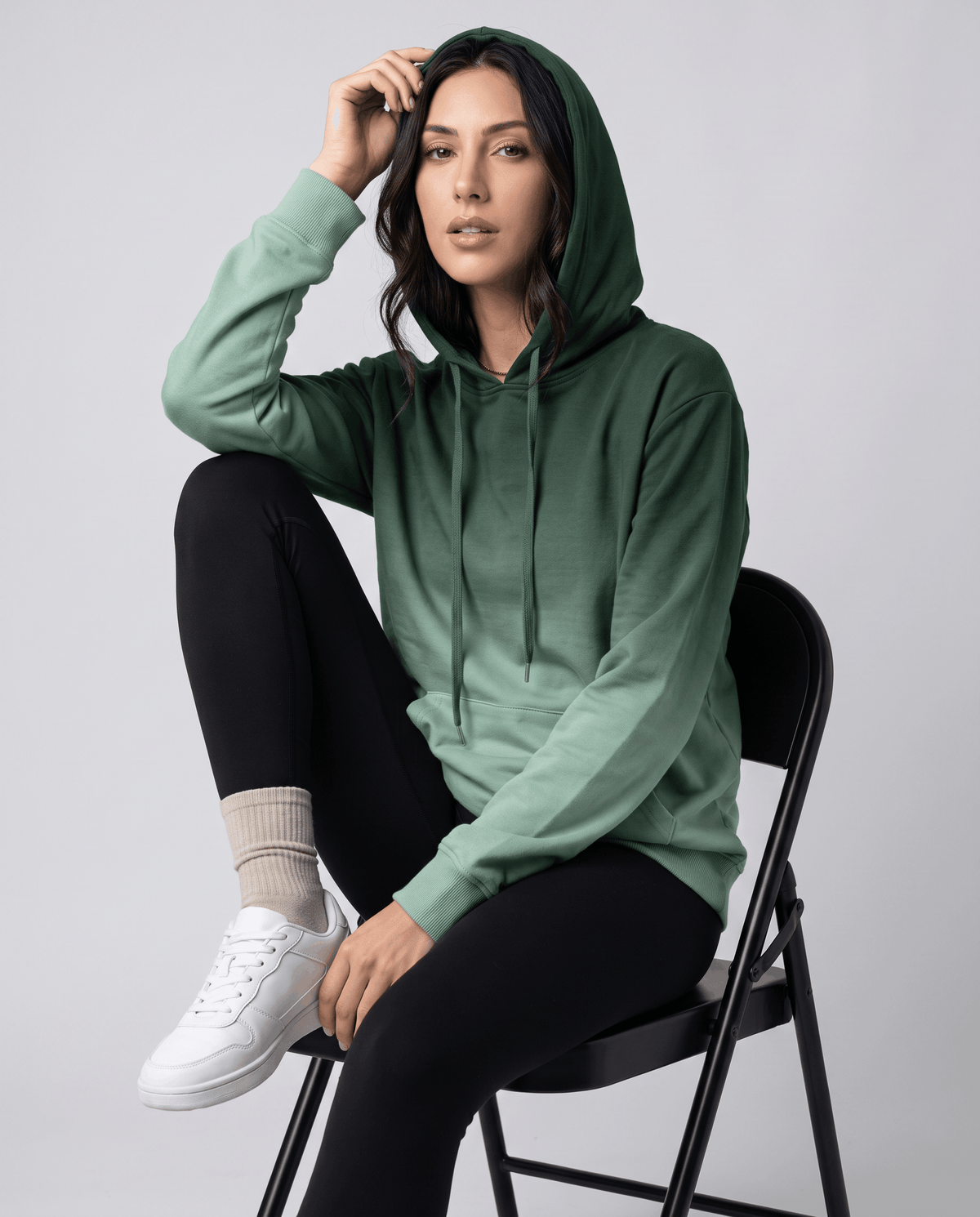 Seated look in the green gradient hoodie with the hood up, black leggings and white sneakers