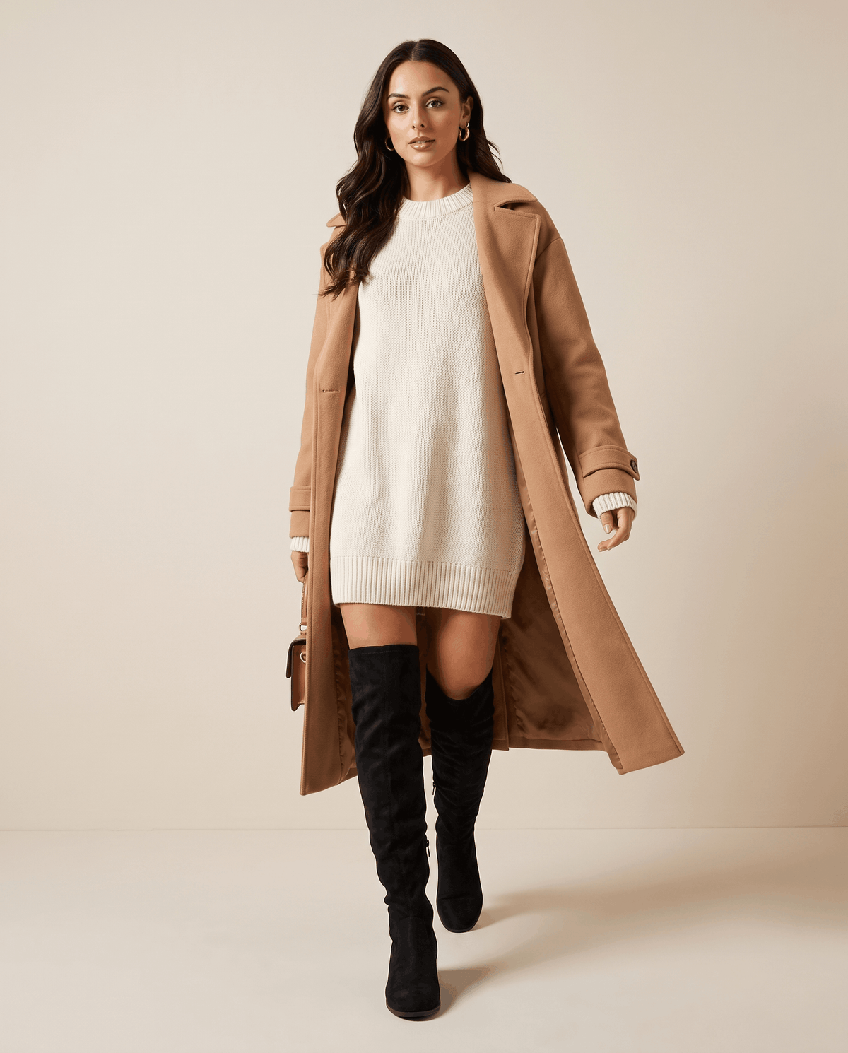 Woman in a camel coat and cream sweater dress styled with black tall boots
