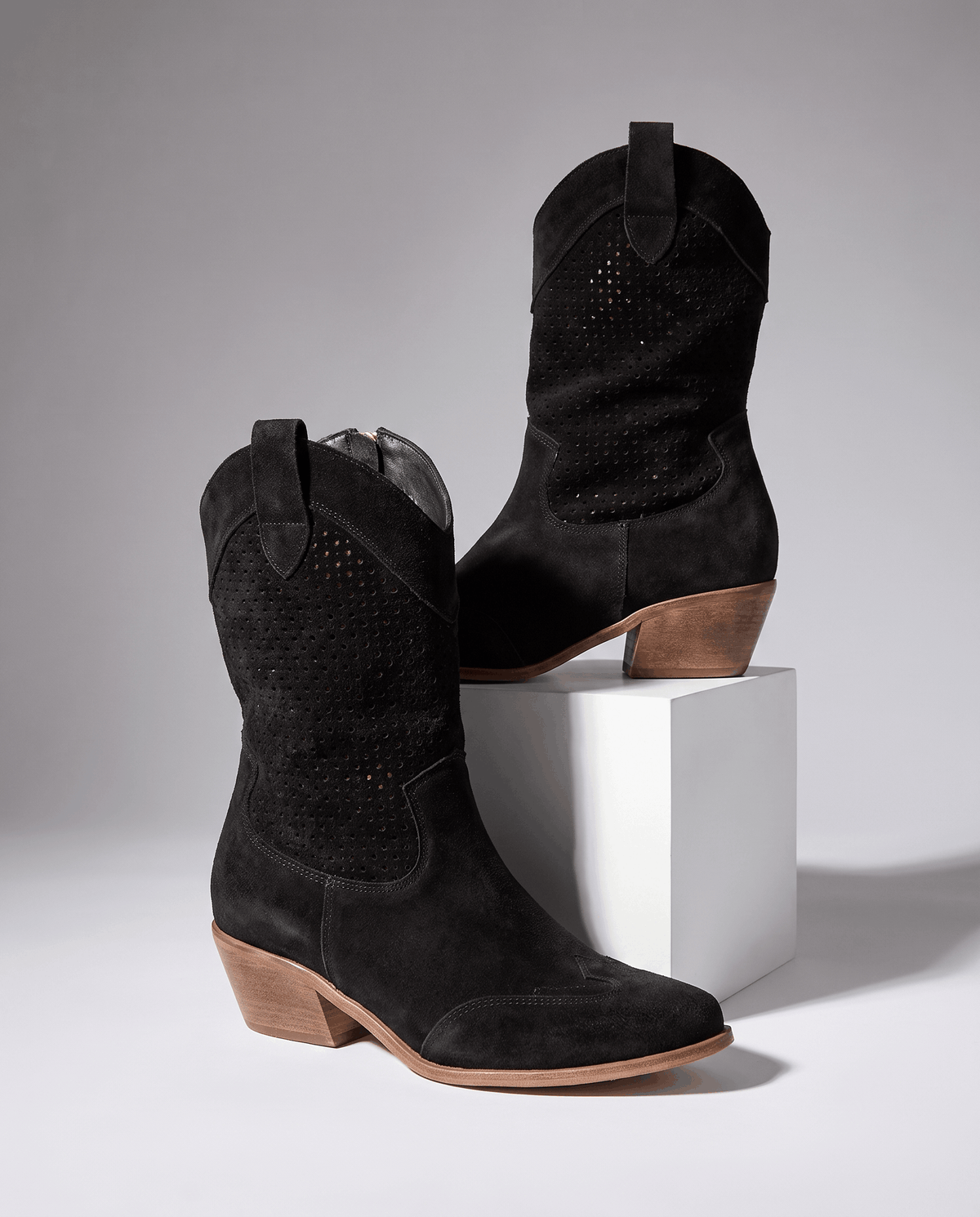 Pair of black perforated western boots displayed on a white pedestal