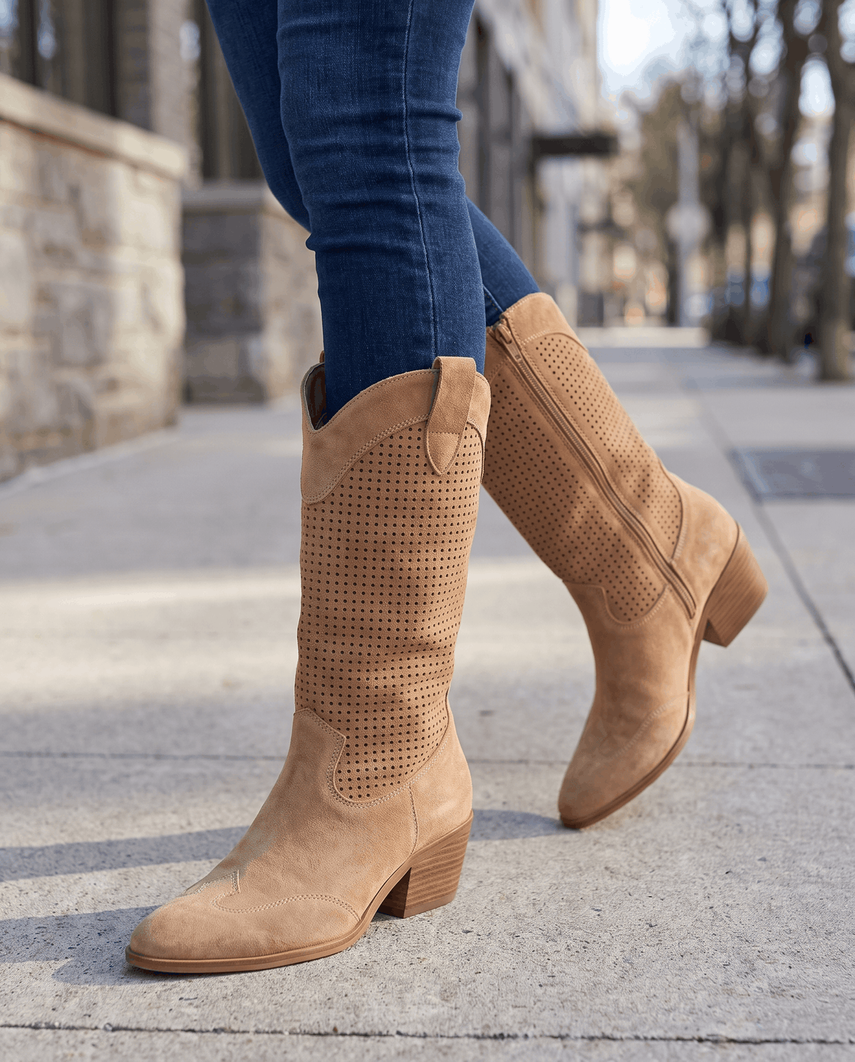 Brown western boots worn walking on a sunny city sidewalk with dark jeans