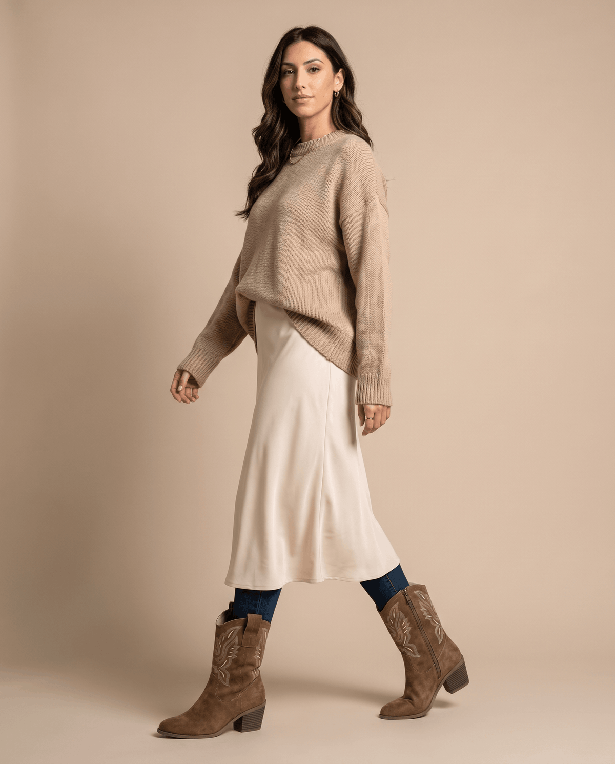 Woman in an oatmeal knit sweater and cream midi skirt walking in camel boots