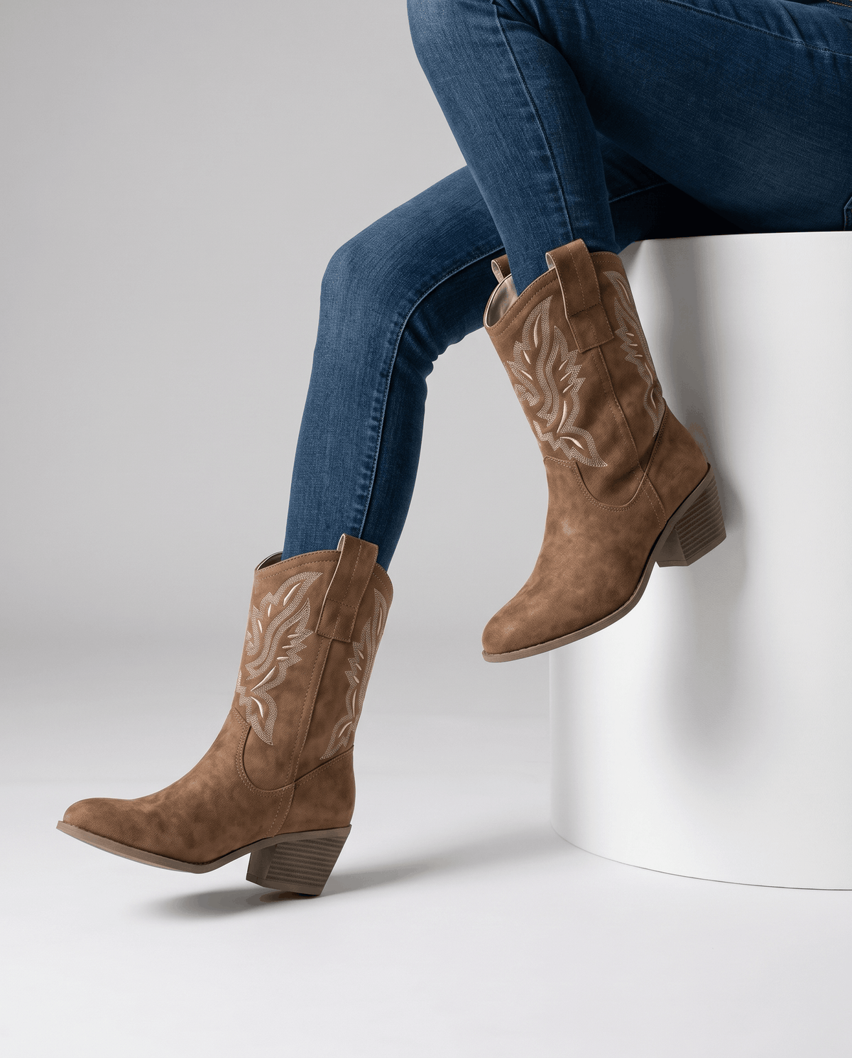 Seated model with blue jeans tucked into camel embroidered boots on a white block