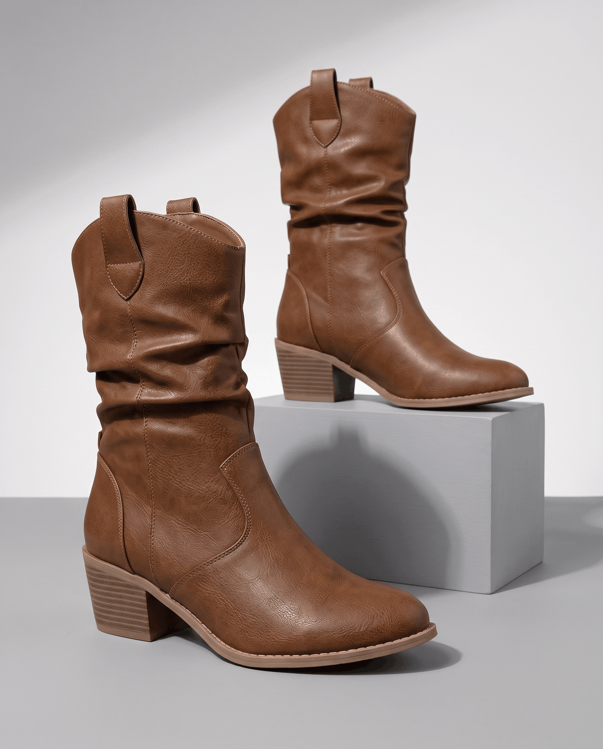 Brown slouchy western boots displayed on a white pedestal against a grey backdrop